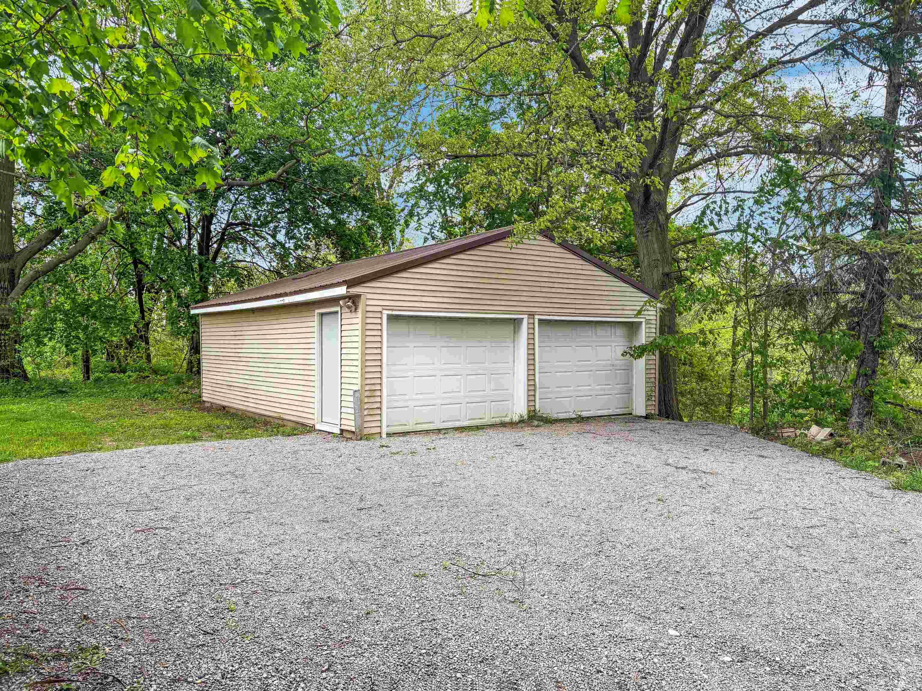 Detached two-car garage with tan horizontal siding, white garage doors, a side entry door, and a brown shingled roof, fronted by a gravel driveway