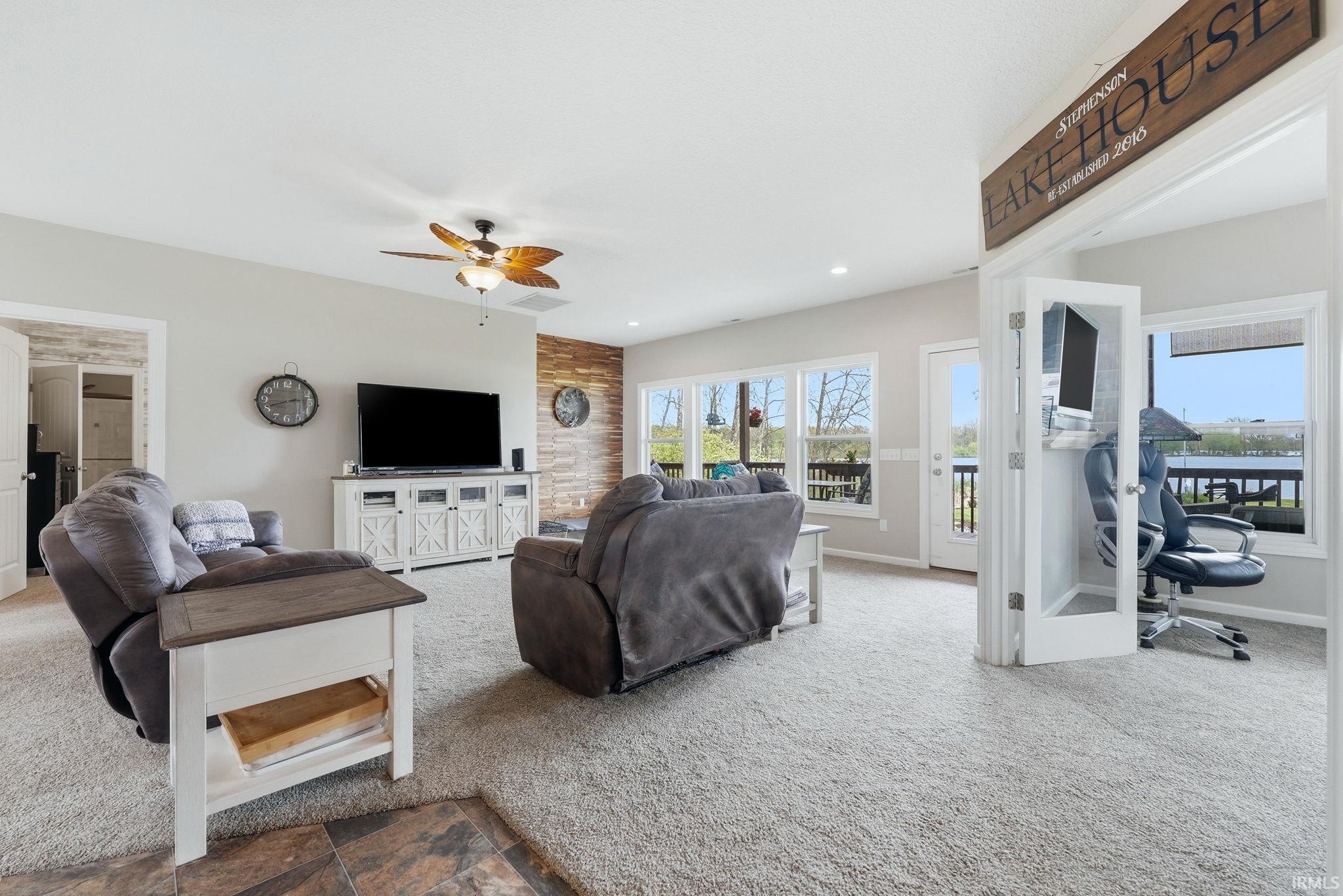 Spacious living area featuring light gray walls, plush carpeting, a ceiling fan with integrated lighting, and an accent wall with stacked wood-finish paneling