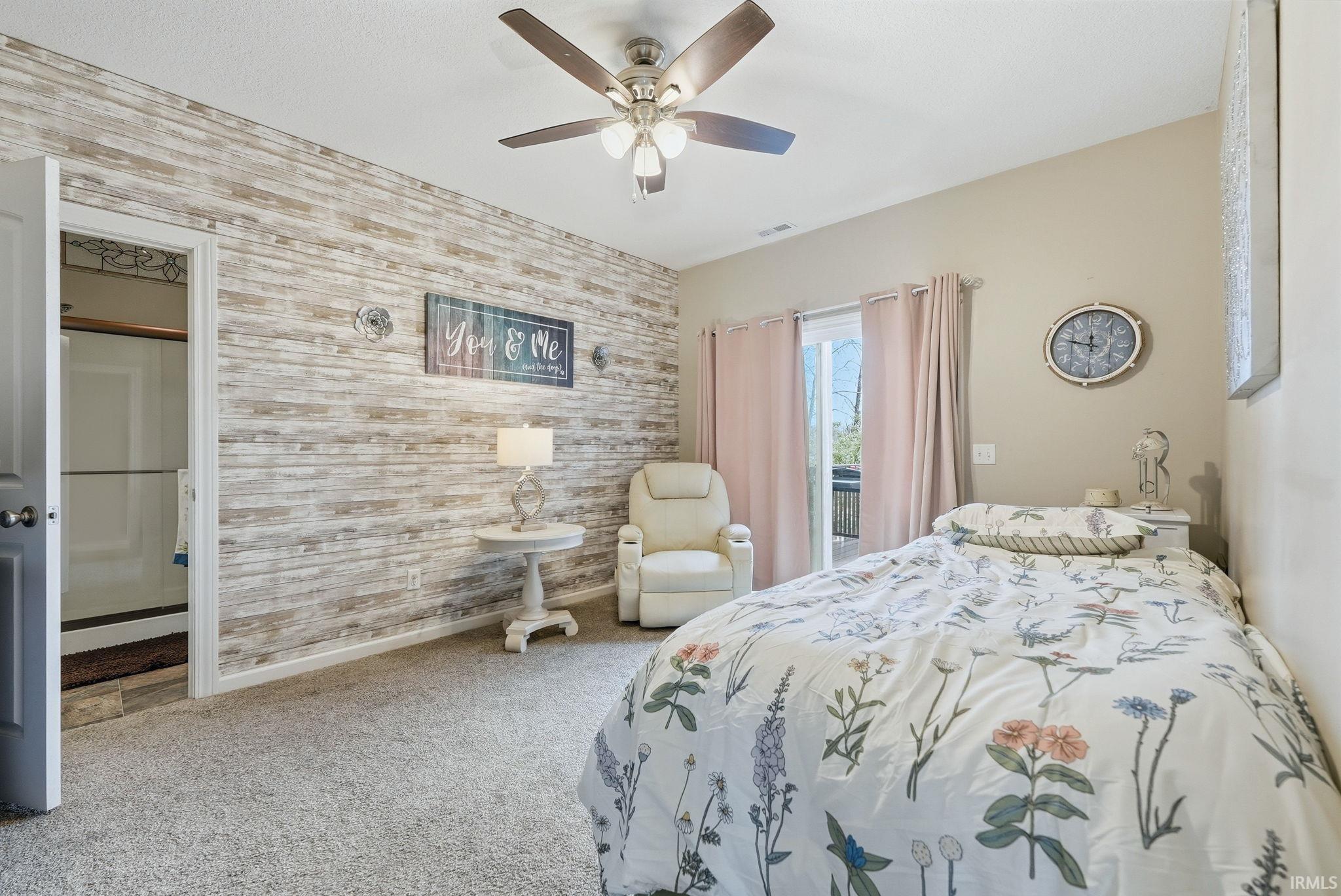 Bedroom featuring a wood-finish accent wall, ceiling fan with integrated lighting, plush carpet flooring, and a sliding glass door to an exterior space