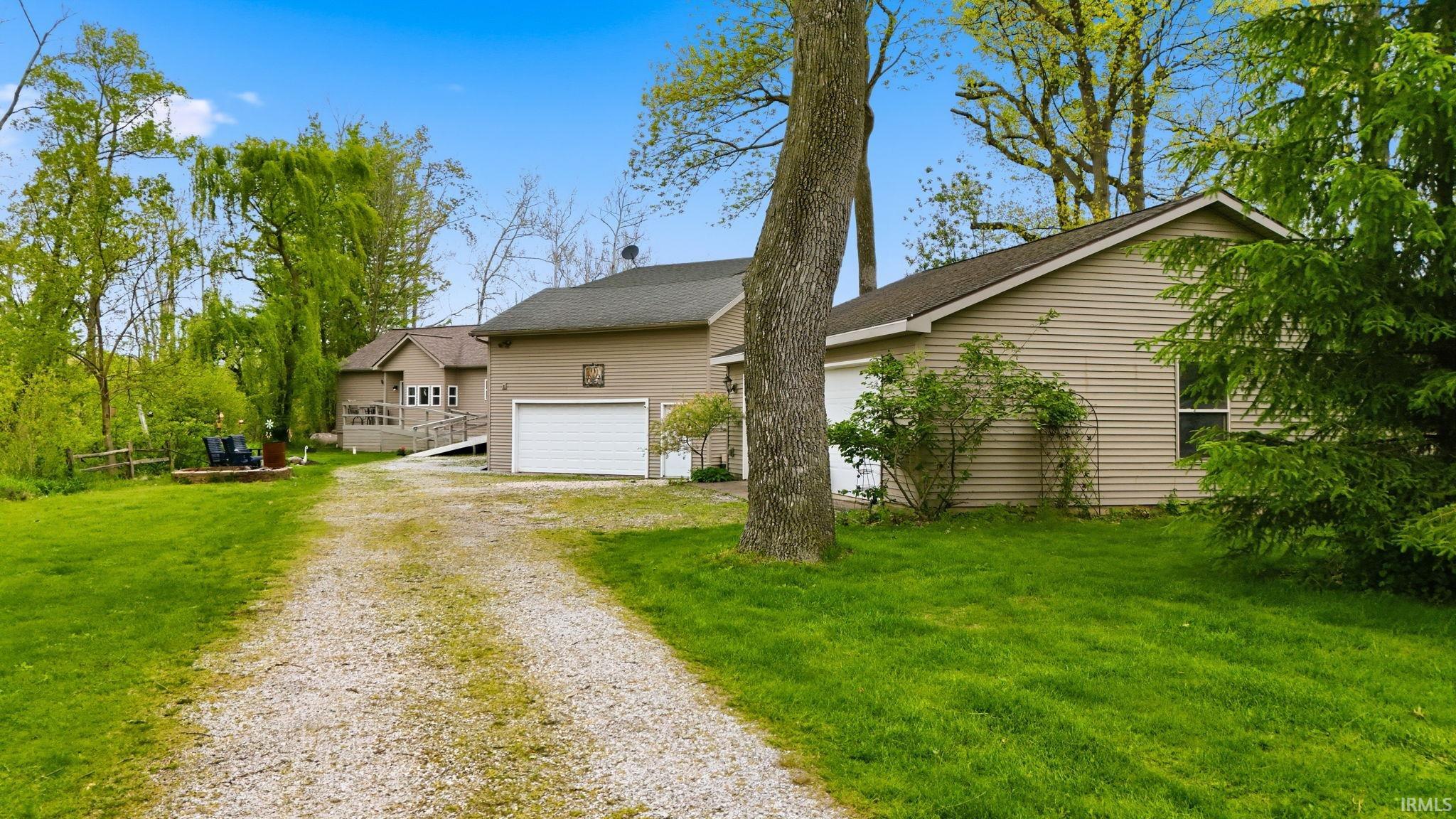 Gravel driveway leading to a residence with tan siding, a white garage door, and a brown shingled roof