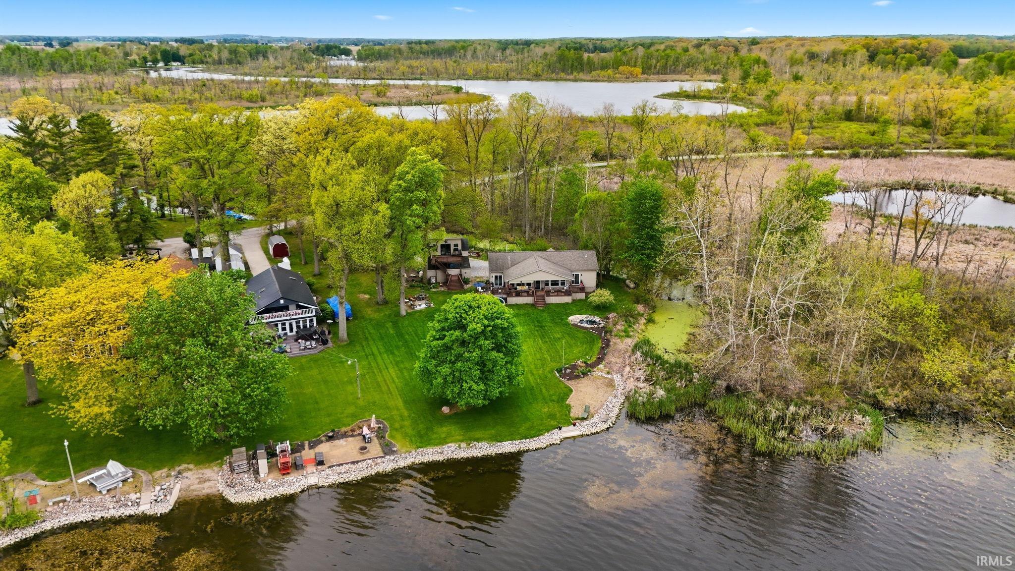 Waterfront property featuring a detached residence, lush green lawn, mature trees, and a stone-lined shoreline