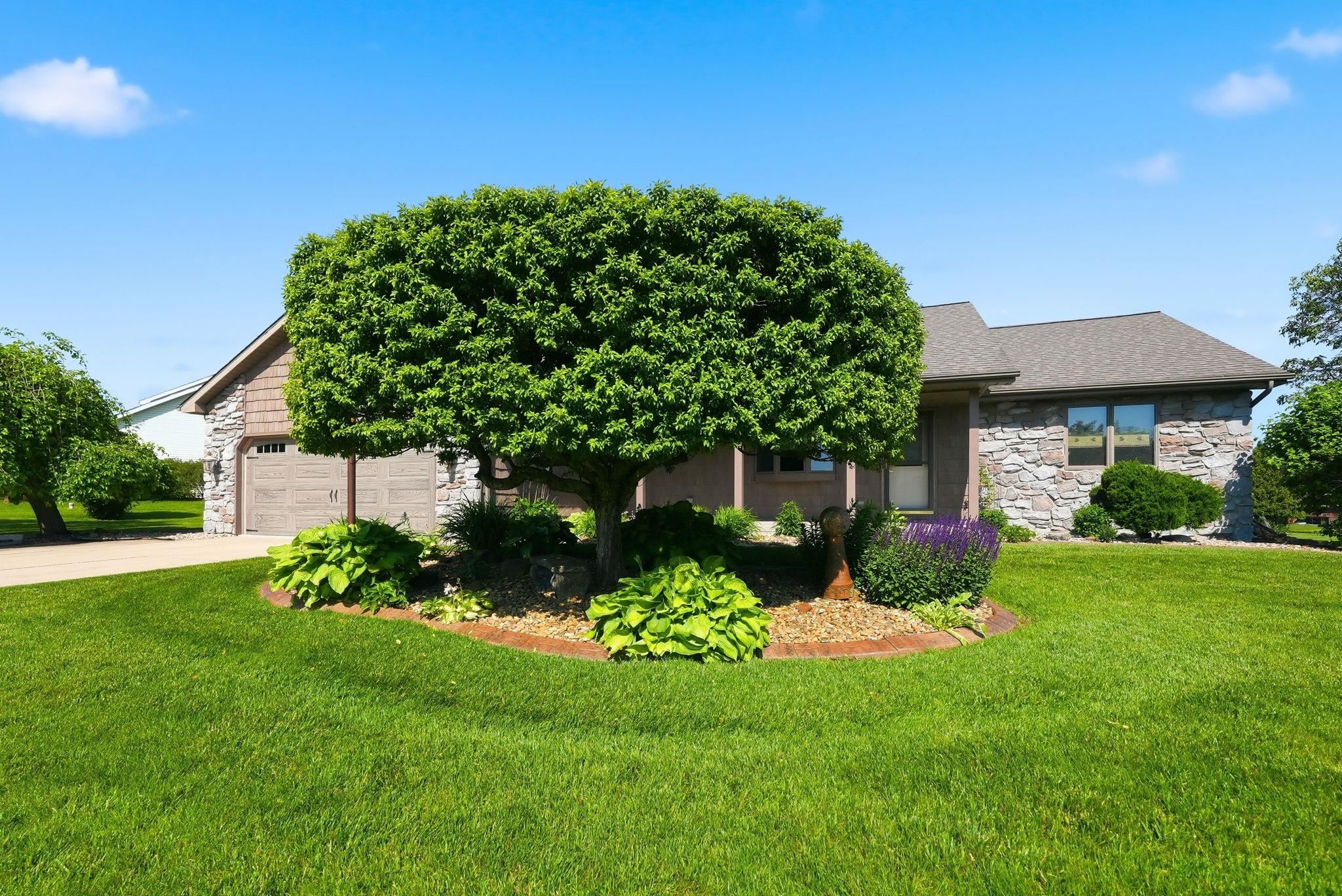 Ranch-style exterior featuring stone veneer siding, a two-car garage with wood-finish doors, and a central landscaped garden bed with mature foliage