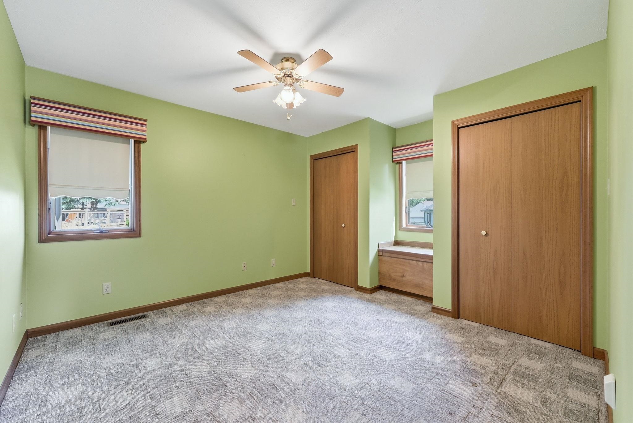 Room featuring patterned carpeting, a ceiling fan with integrated lighting, two windows with blinds and valances, and two wood-finish bi-fold closet doors