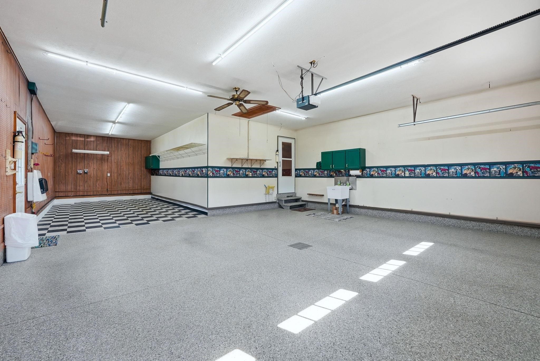 Spacious garage featuring a durable speckled epoxy floor, wood-paneled wall, overhead lighting, ceiling fan, and built-in utility sink with cabinetry
