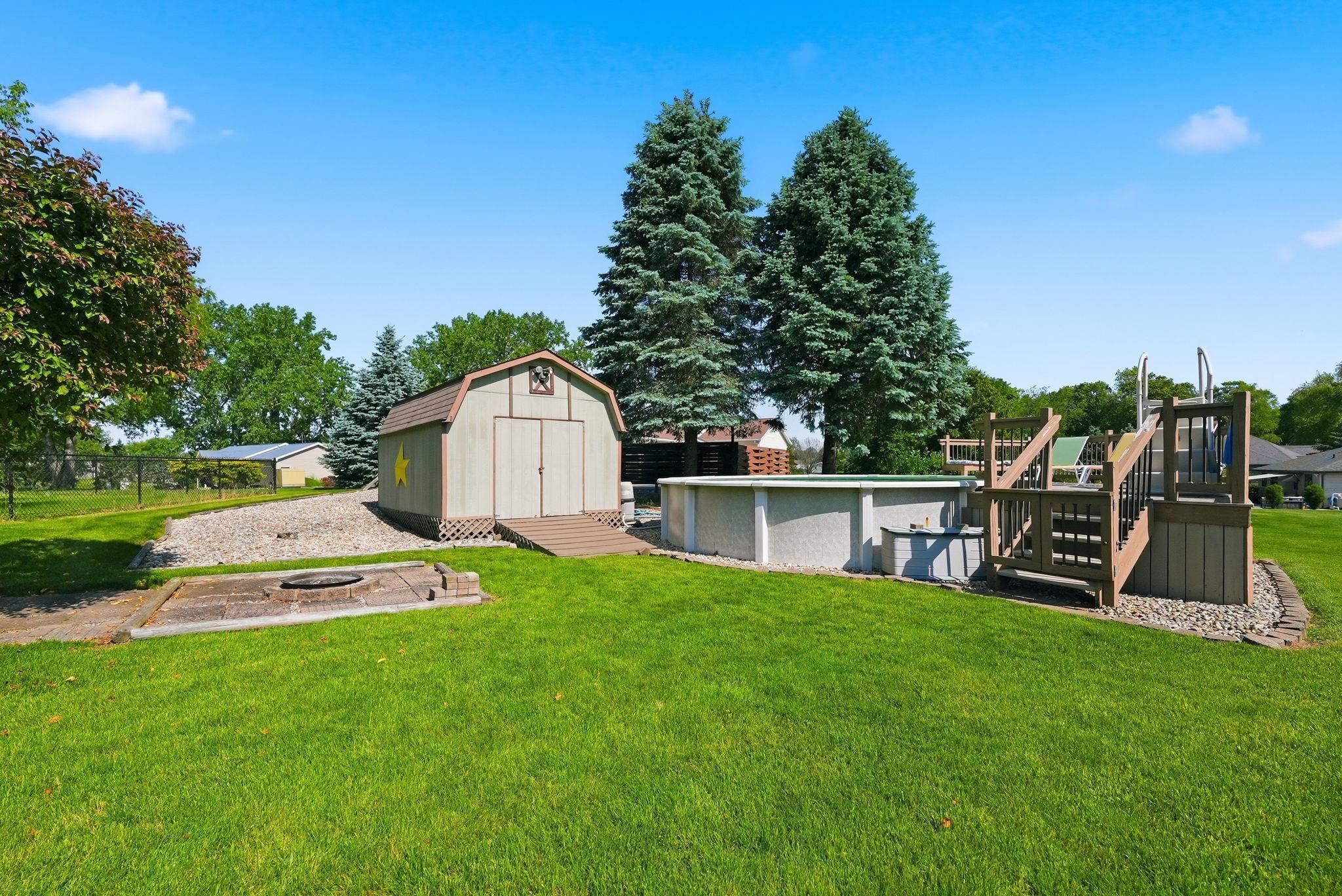 Expansive backyard featuring a lush lawn, above-ground pool with an accompanying wooden deck, a storage shed with a ramp, and a fire pit area
