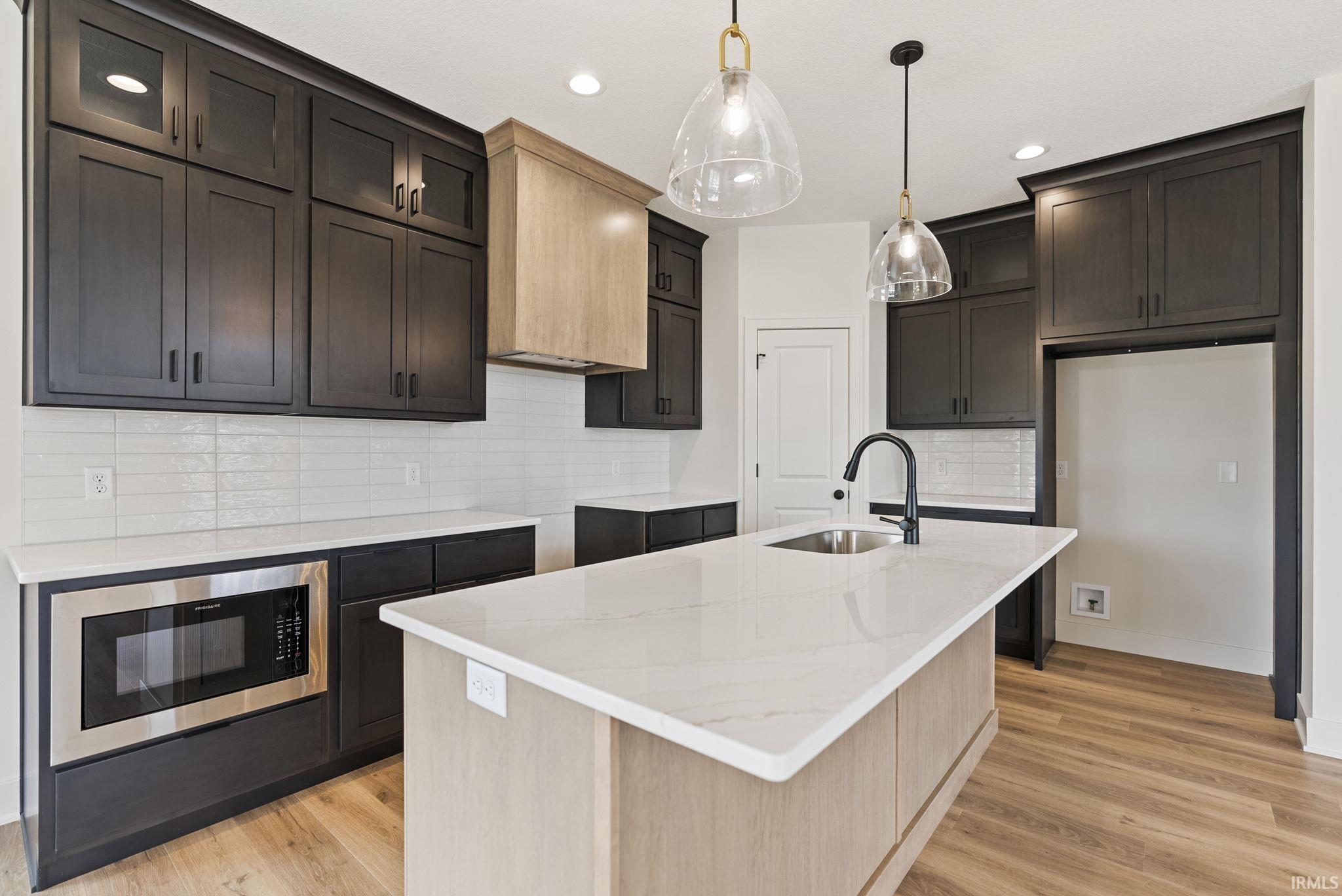 Kitchen featuring a large island with an undermount sink and matte black faucet, dark cabinetry, light countertops, and a subway tile backsplash