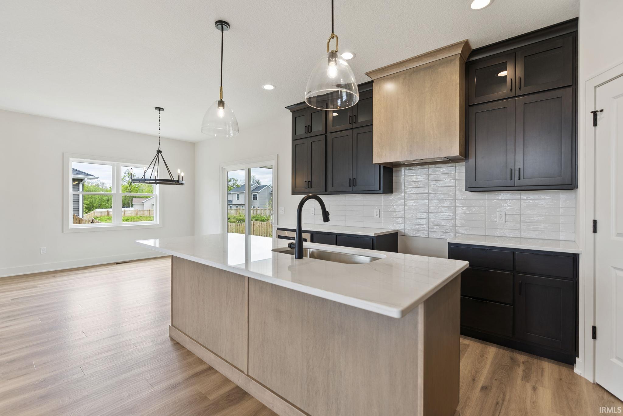 Contemporary kitchen featuring a center island with an integrated sink, dark cabinetry, light wood-finish flooring, white subway tile backsplash, and a light wood range hood