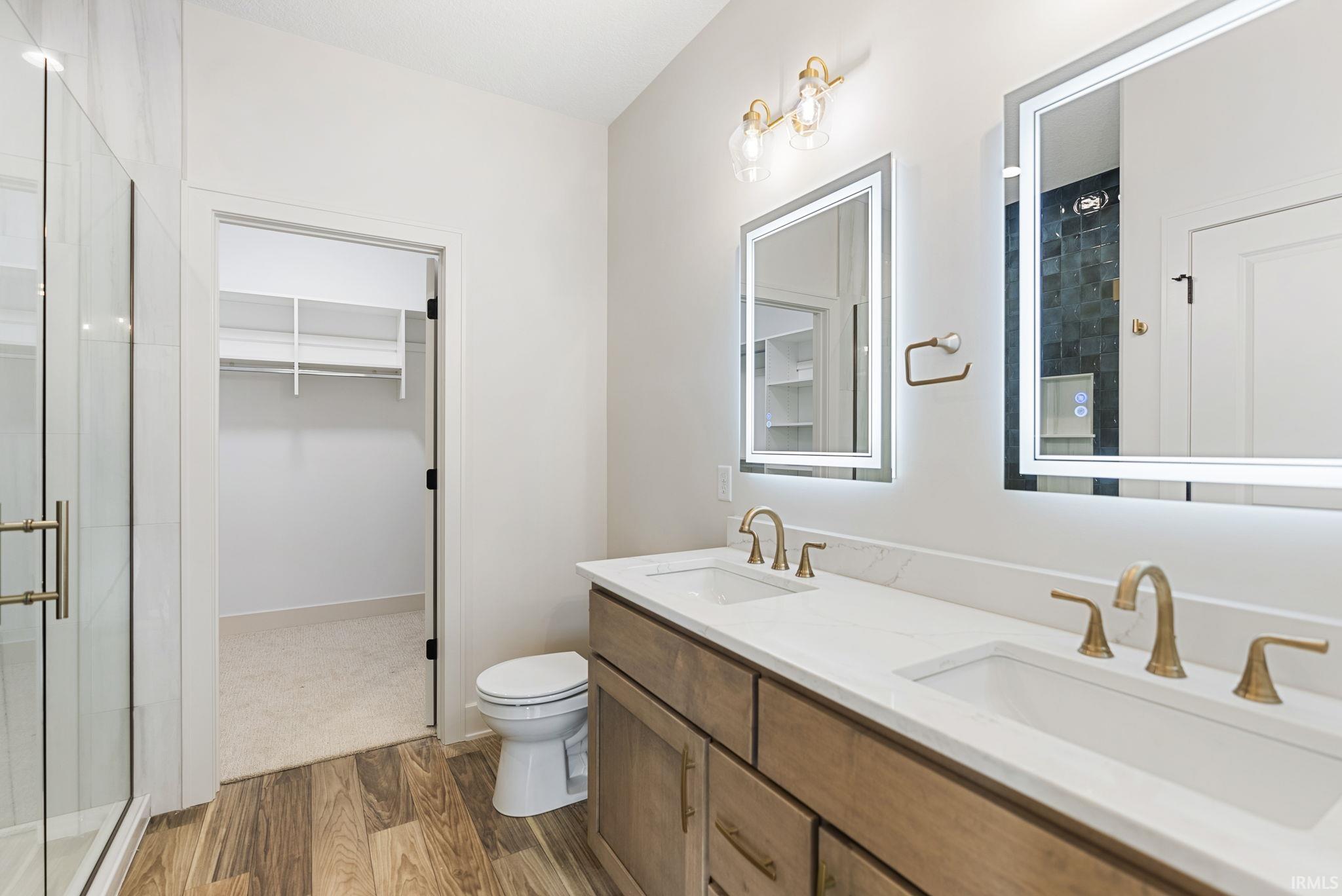 Bathroom featuring a dual vanity with undermount sinks and brass-finish faucets, illuminated mirrors, a glass-enclosed shower, and wood-finish flooring