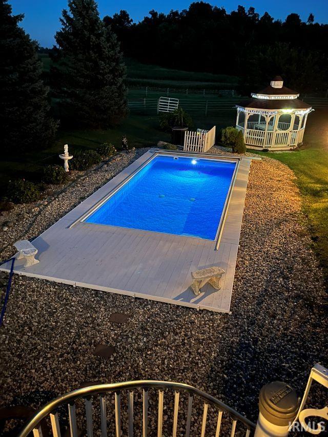 Illuminated inground pool with a white deck surround, featuring a white gazebo and a gravel landscape