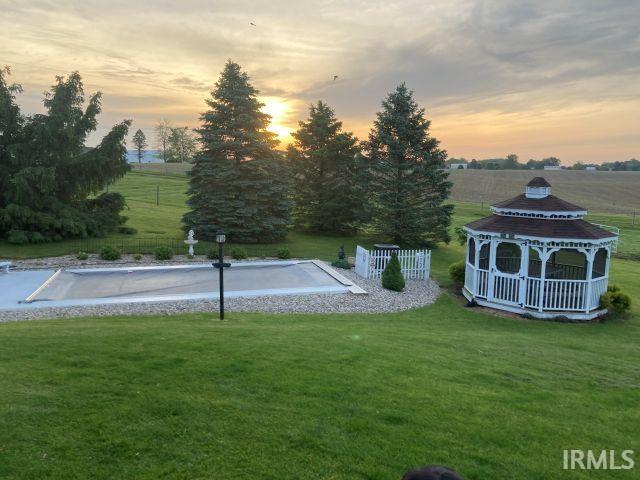 Expansive backyard featuring a white gazebo with a dark shingle roof, a fenced swimming pool area with a gravel border, mature evergreen trees, and a manicured lawn