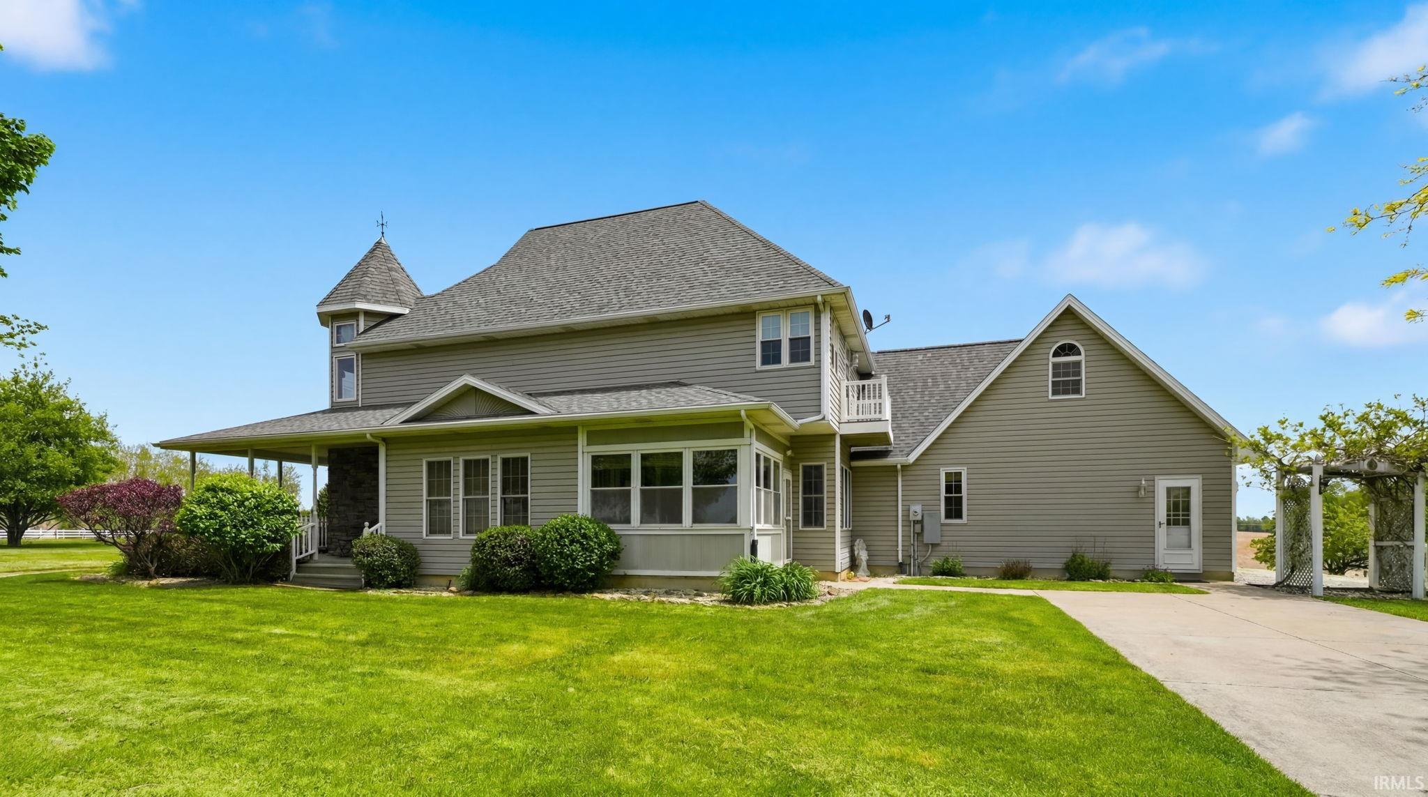 Exterior featuring light gray siding, a prominent turret, a full-length covered porch, an enclosed sunroom, and a paved driveway leading to a rear entrance