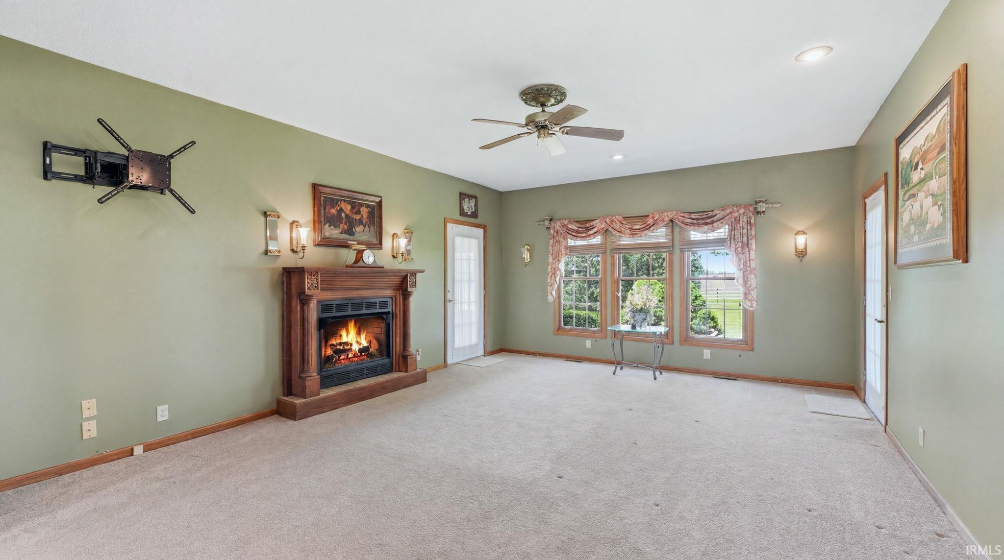Spacious room featuring a decorative wood mantel fireplace, light beige carpeting, multiple windows, and a ceiling fan