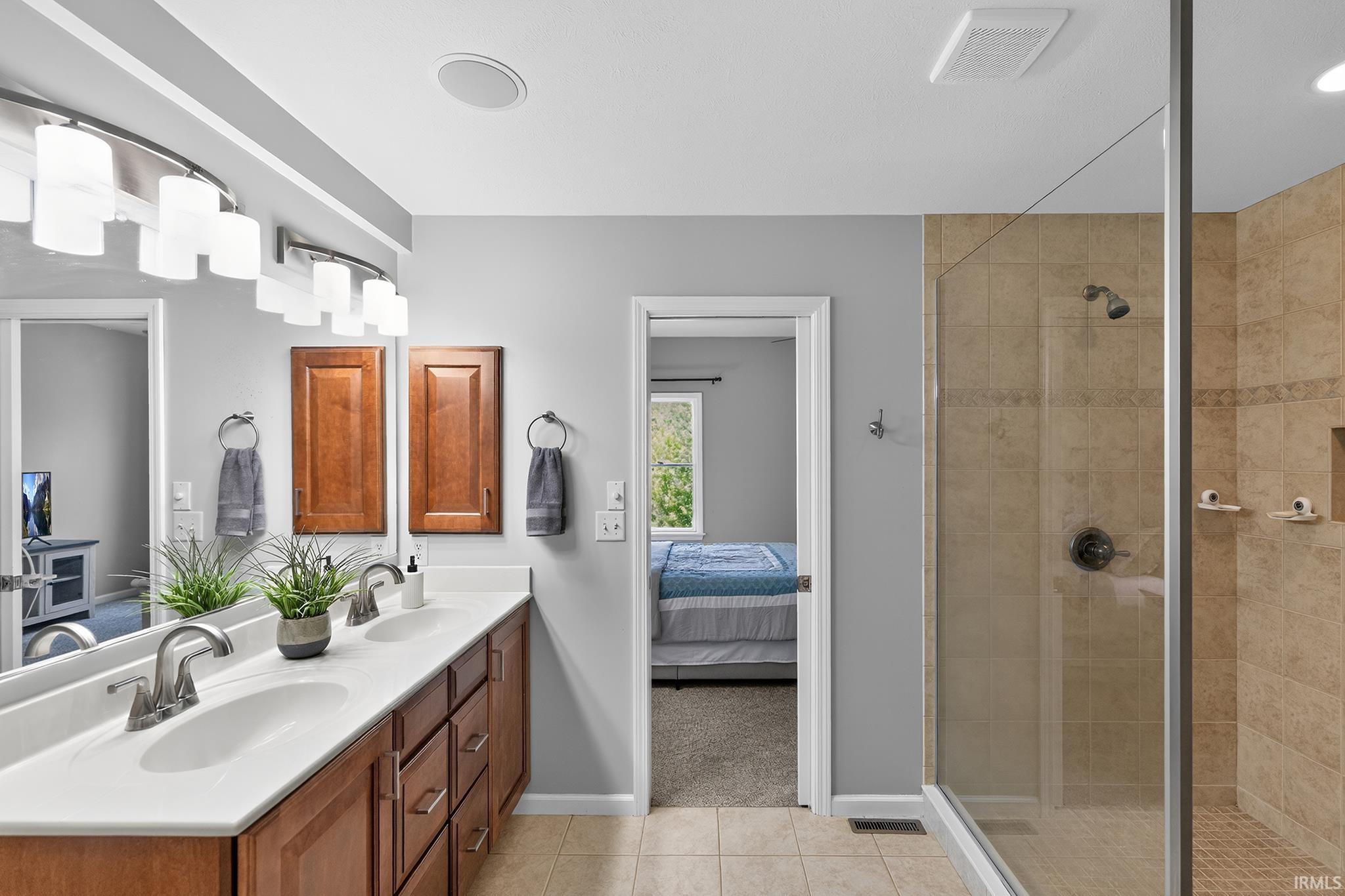 Bathroom featuring a dual-sink vanity with wood-finish cabinetry, a large frameless mirror, and a tiled walk-in shower with a glass enclosure
