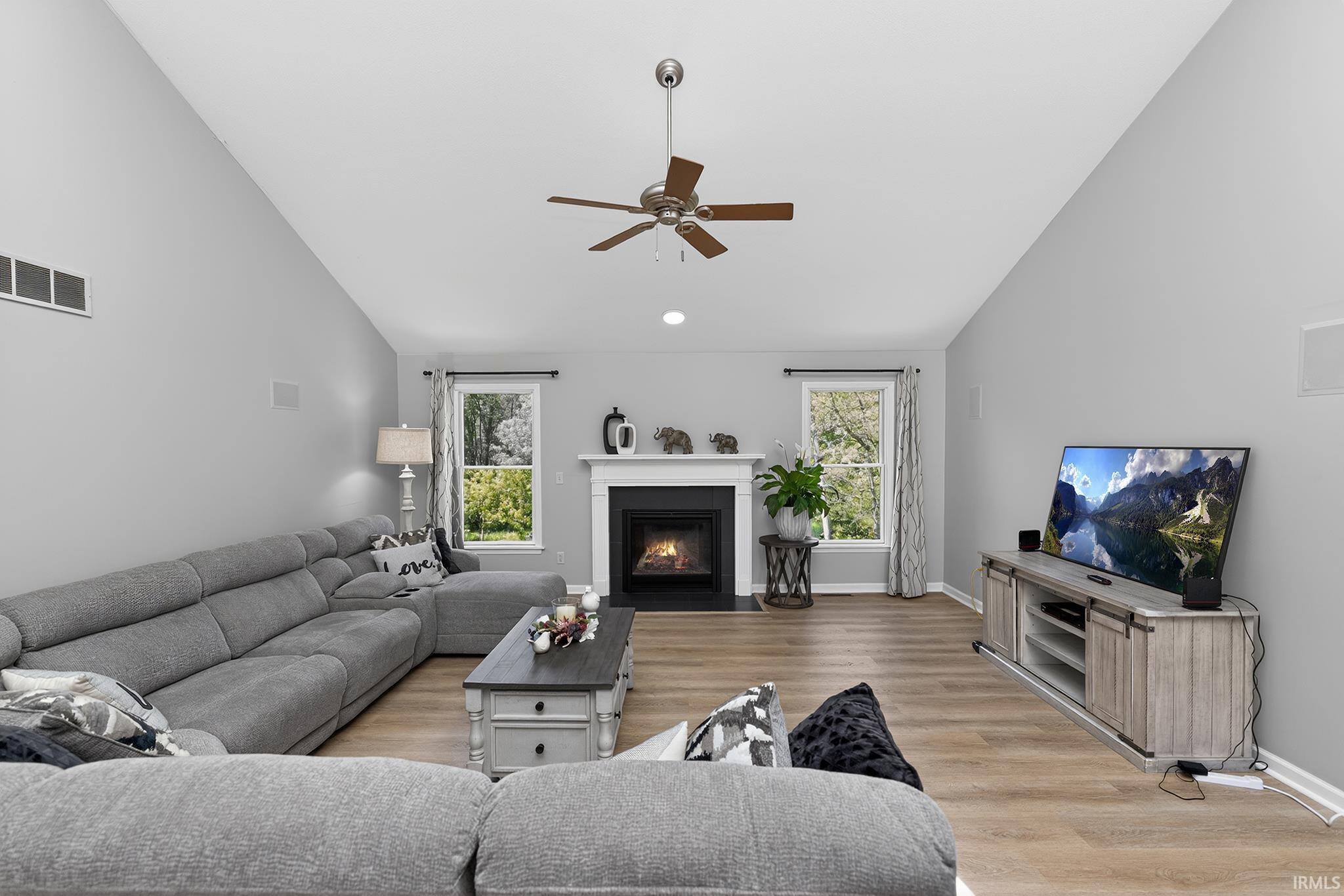 Spacious living area featuring a vaulted ceiling with a ceiling fan, a fireplace with a white mantel, and wood-finish flooring