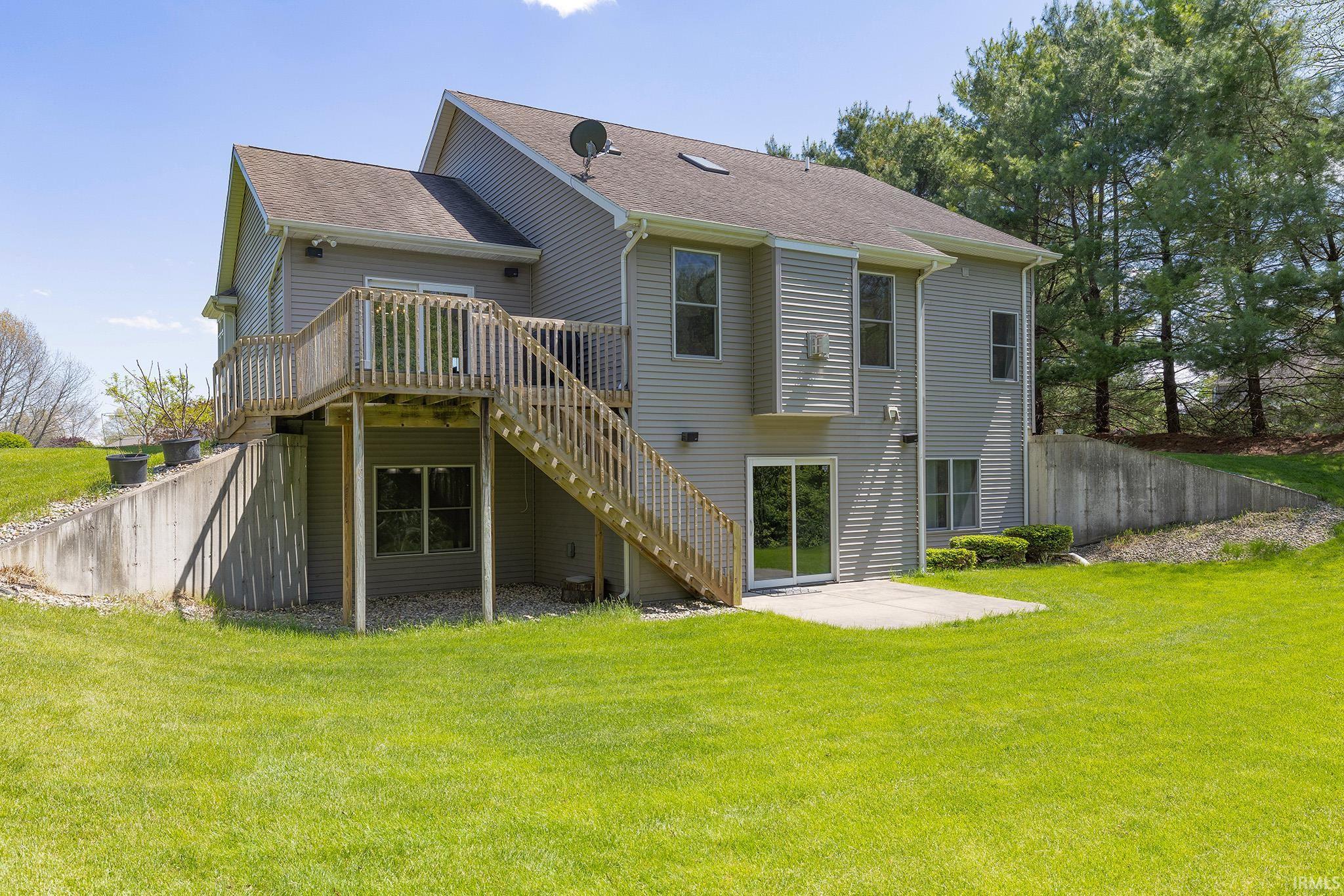 Expansive rear facade featuring a wooden deck with staircase access to the lawn, a ground-level concrete patio, and mature trees bordering the property