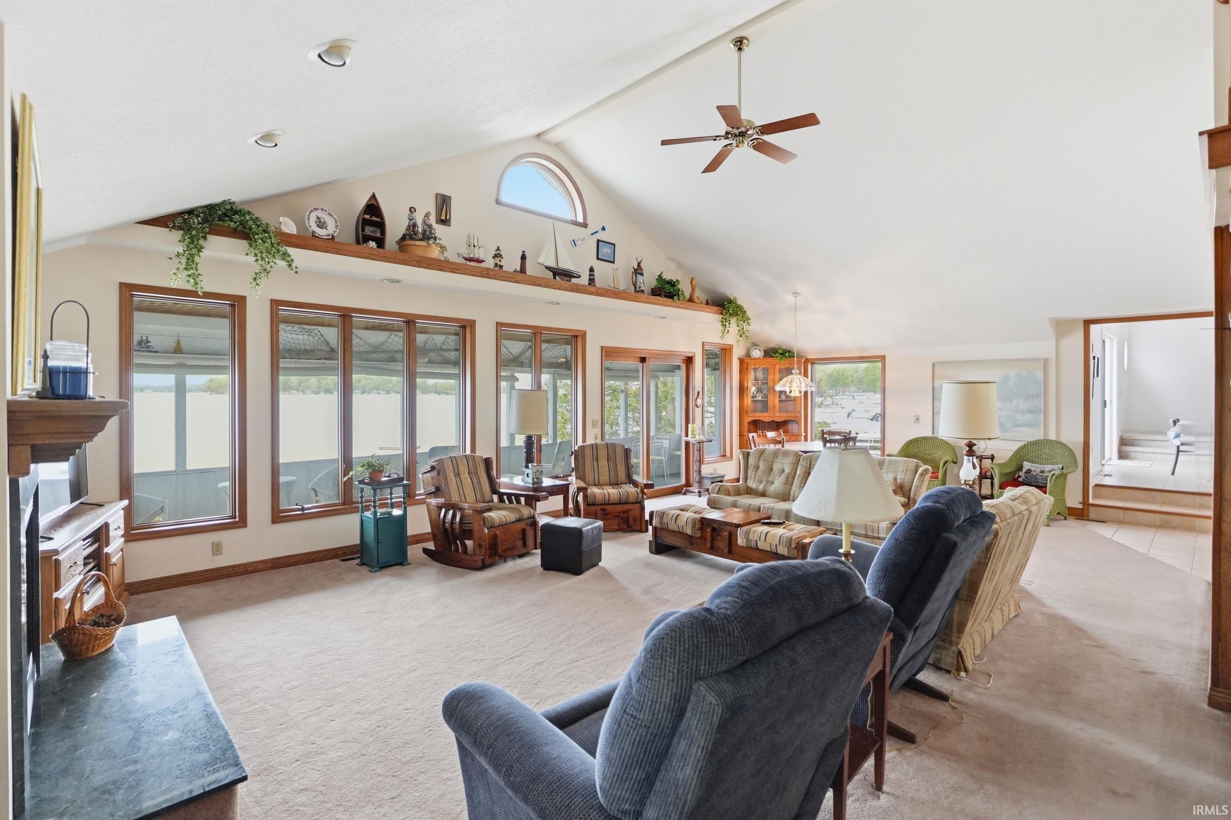 Spacious living area featuring a vaulted ceiling with a ceiling fan, a prominent fireplace with a dark stone hearth, and multiple large windows with wood-finish frames