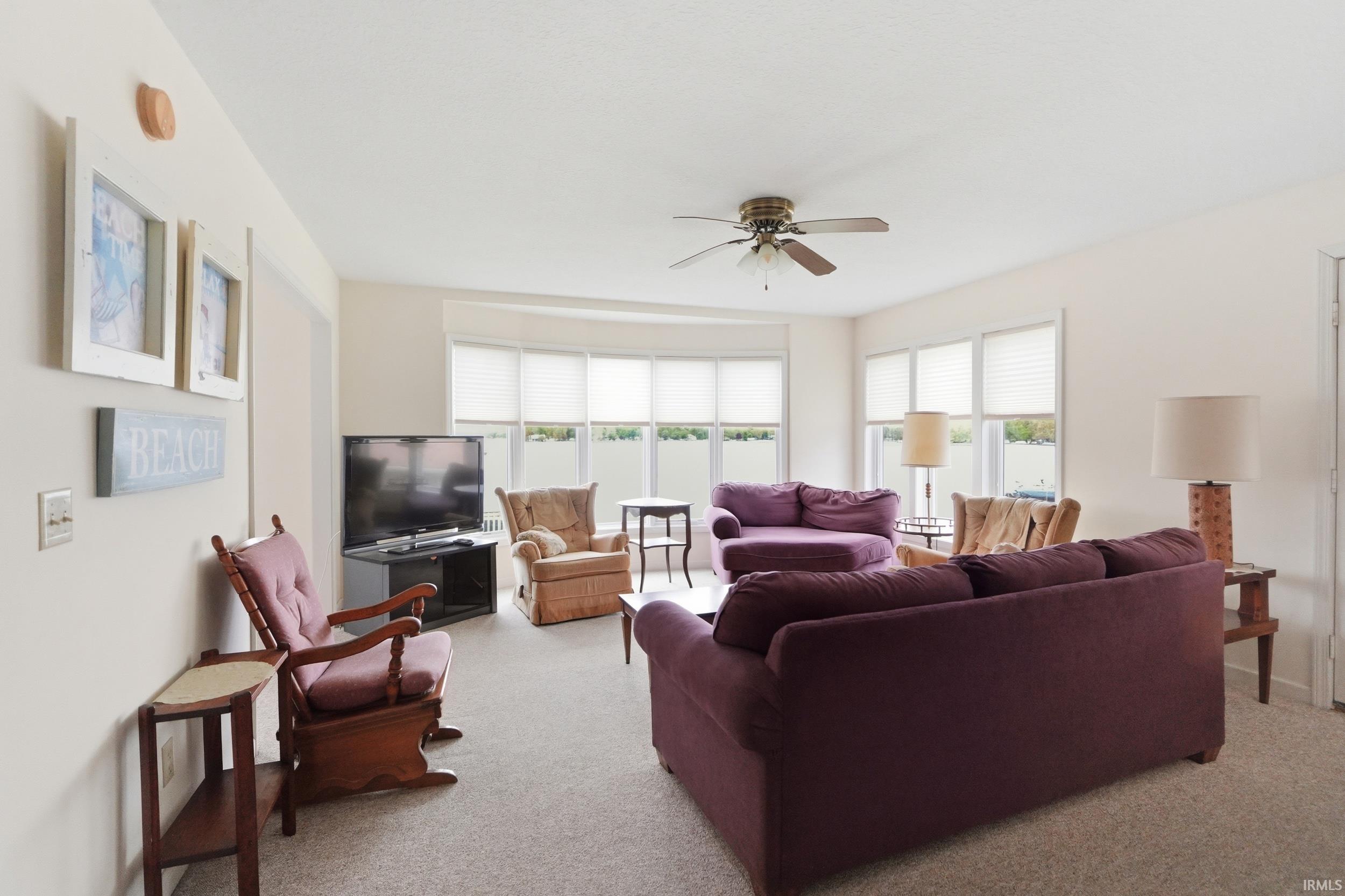 Carpeted living space featuring a bay window with multiple vertical blinds, a ceiling fan with integrated lighting, and neutral wall tones