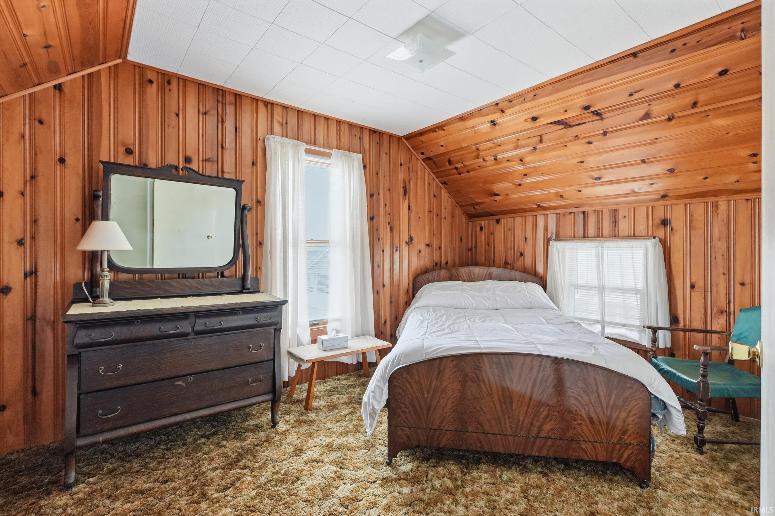 Sloped ceiling room featuring wood paneling, textured carpet flooring, and a single window with white curtains