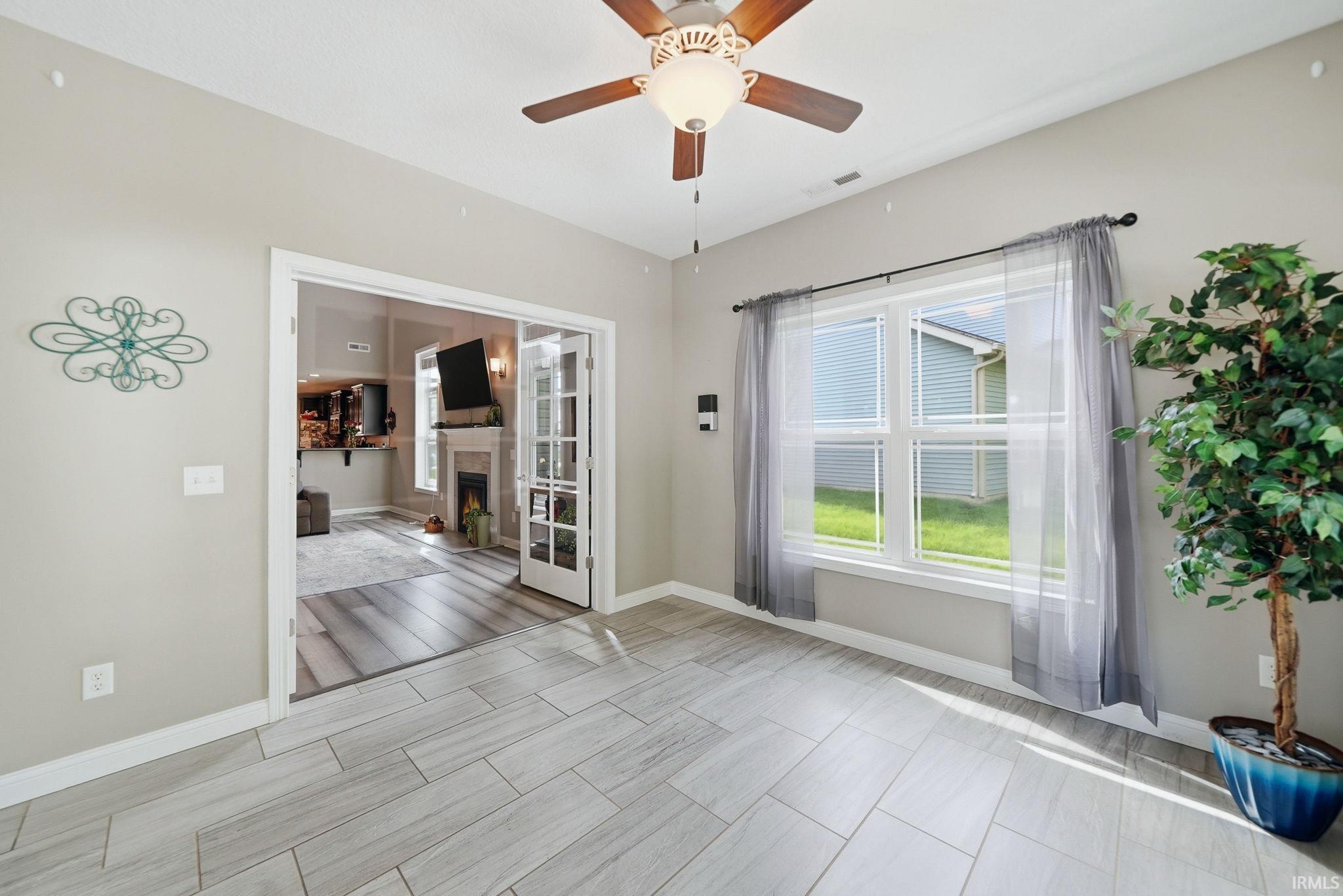 Light-toned wood-look tile flooring in a spacious room featuring a ceiling fan with integrated lighting, a large window with sheer curtains, and a doorway with French doors opening to another room with wood-finish flooring and a fireplace