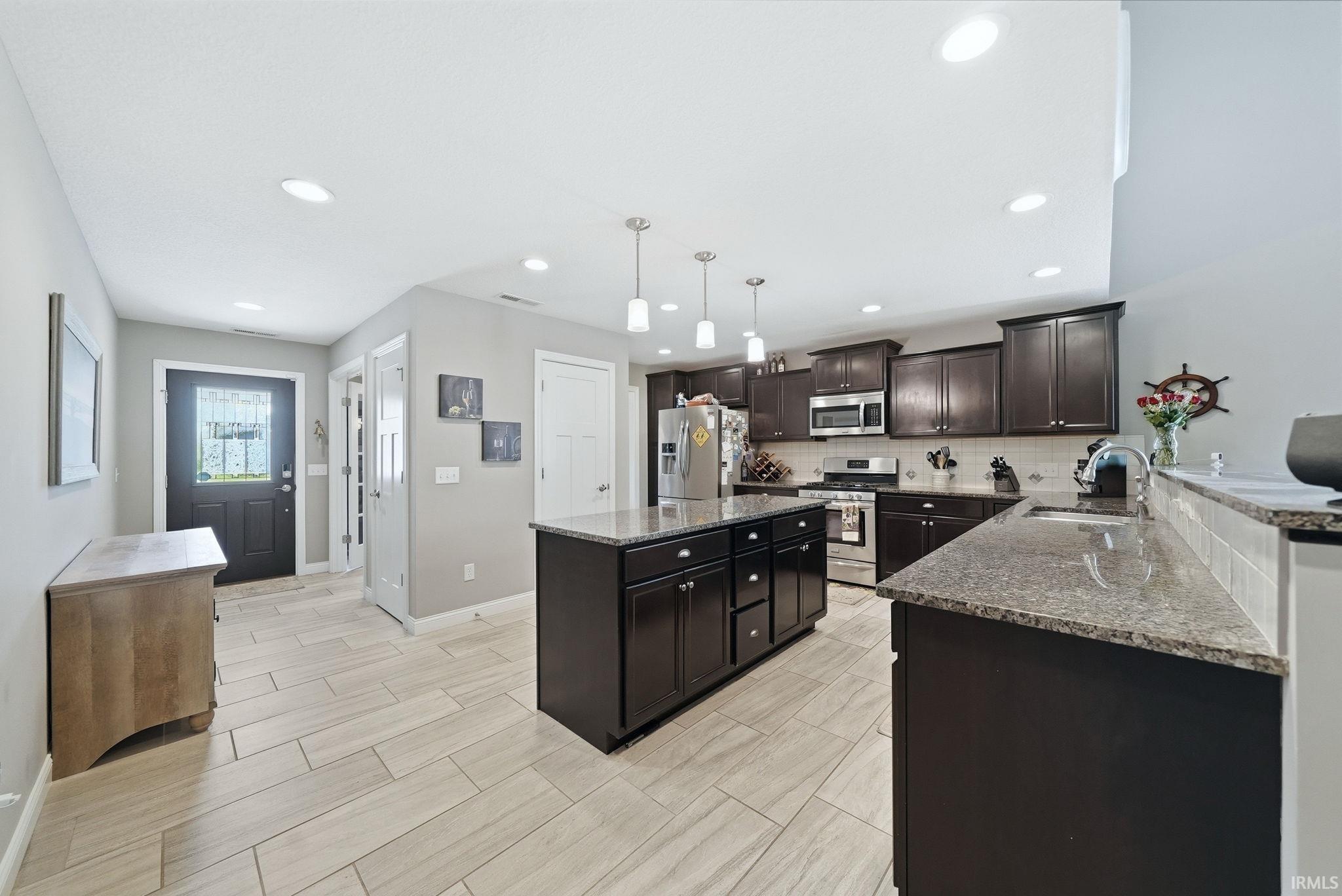 Open-concept kitchen featuring dark wood cabinetry, a central island with pendant lighting, stone countertops, stainless steel appliances, and wood-finish tile flooring