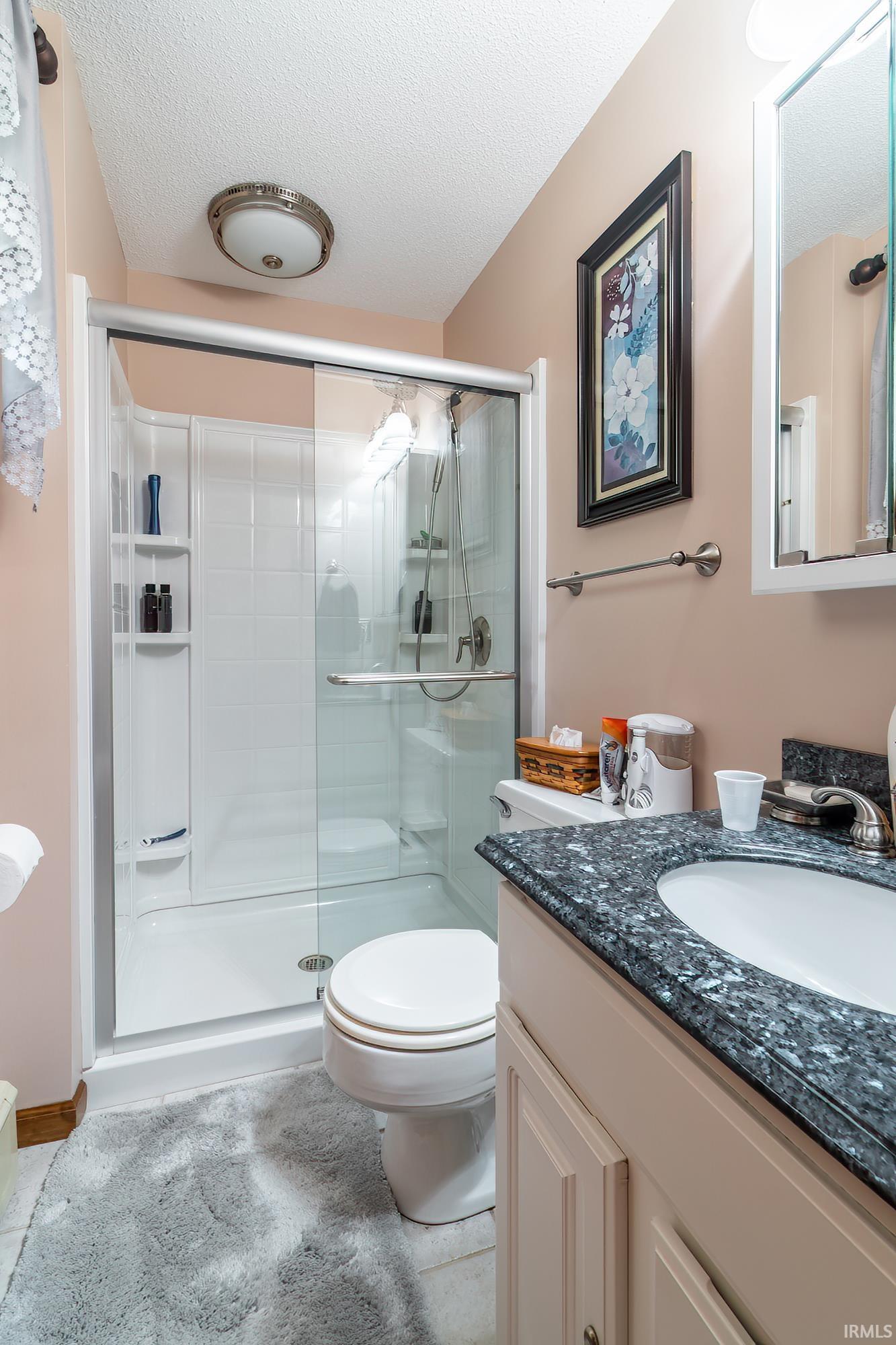 Bathroom featuring a glass-enclosed shower with white surround, a commode, and a vanity with a dark speckled countertop and integrated sink