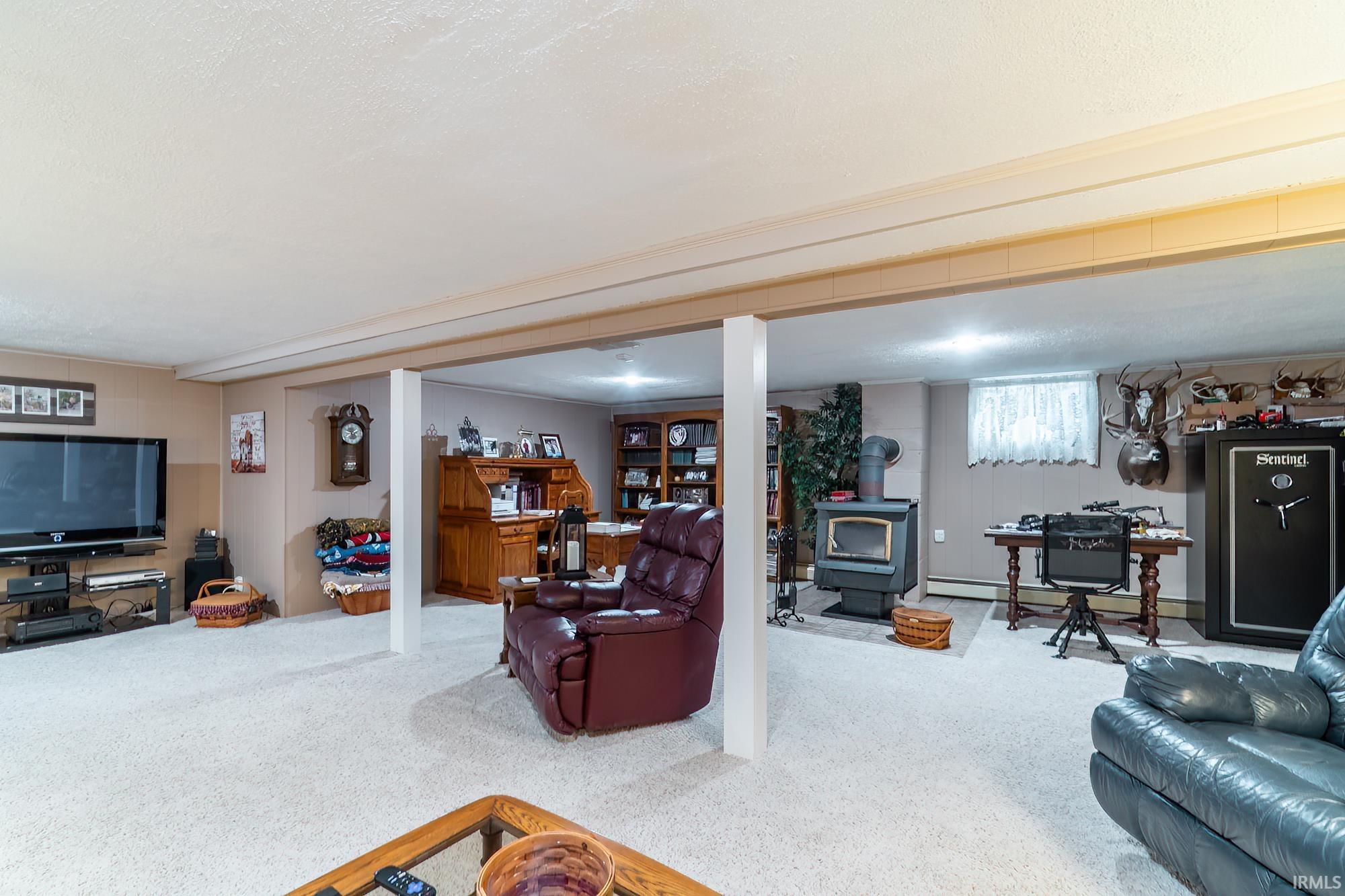 Finished basement featuring plush carpeting, exposed ceiling beams, a built-in wood-burning stove with a stone hearth, recessed lighting, and a window with white blinds