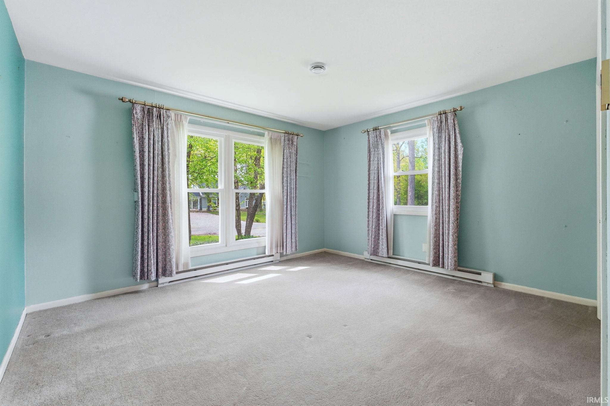 Light-filled room featuring two large windows, neutral carpeting, and baseboard heating