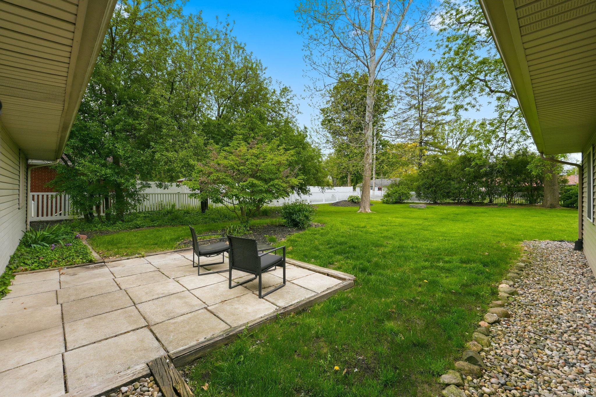 Expansive backyard featuring a stone paver patio, a manicured lawn, established trees, and a white picket fence