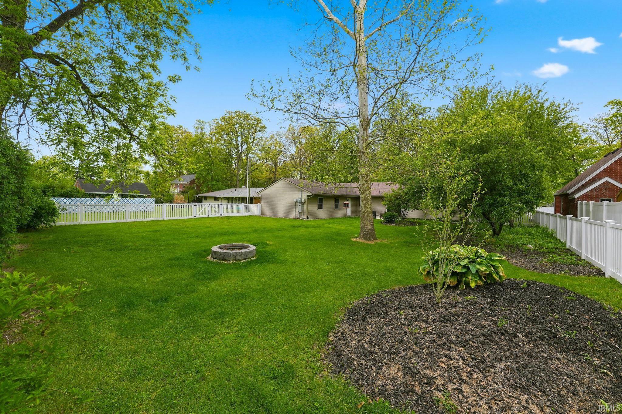 Expansive green lawn with a built-in fire pit, surrounded by mature trees and a white picket fence