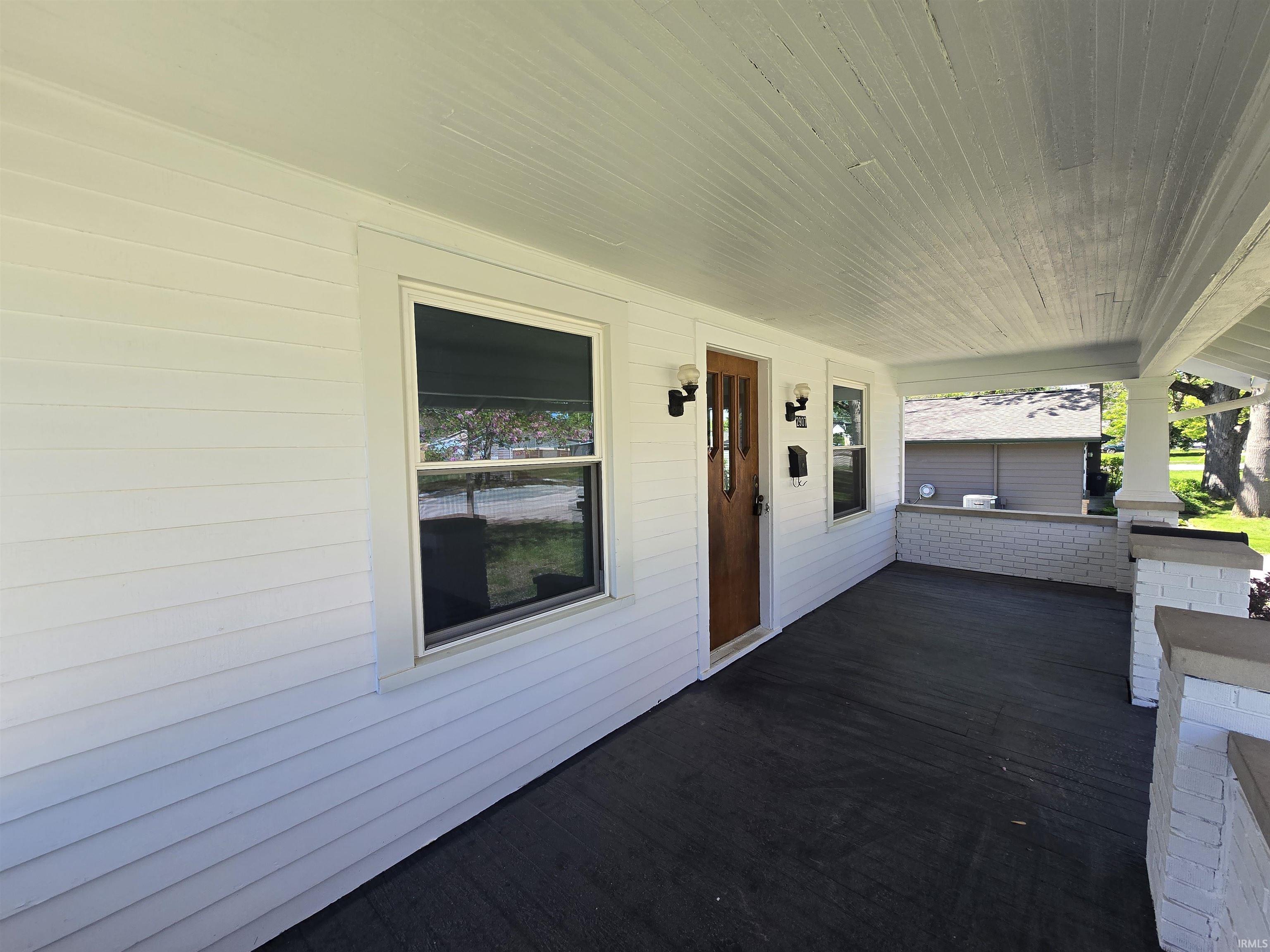 Covered front porch featuring white horizontal siding, a natural wood-finish front door, white painted brick accents, and dark flooring