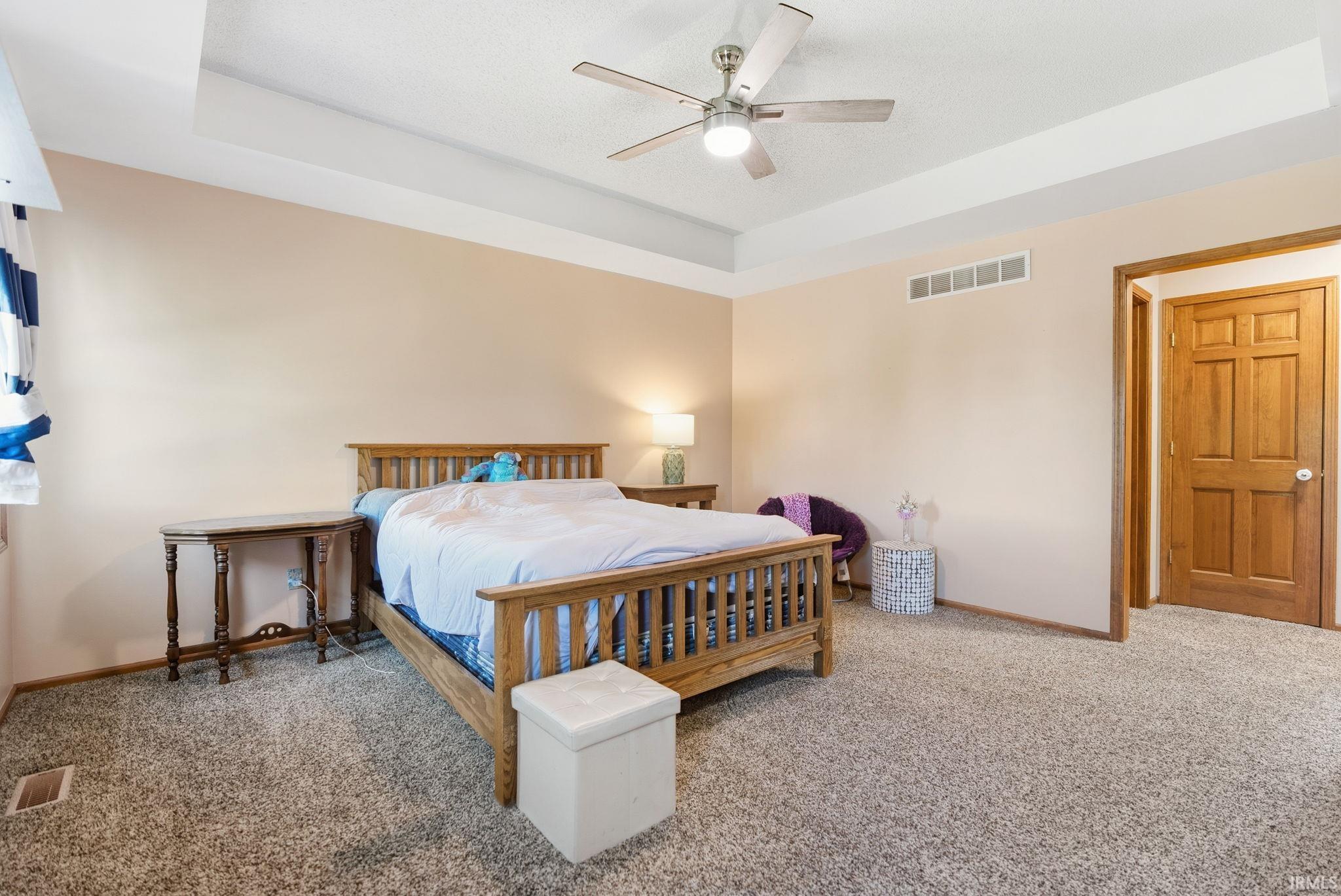 Spacious bedroom featuring a tray ceiling with a ceiling fan, neutral wall tones, and plush carpet flooring