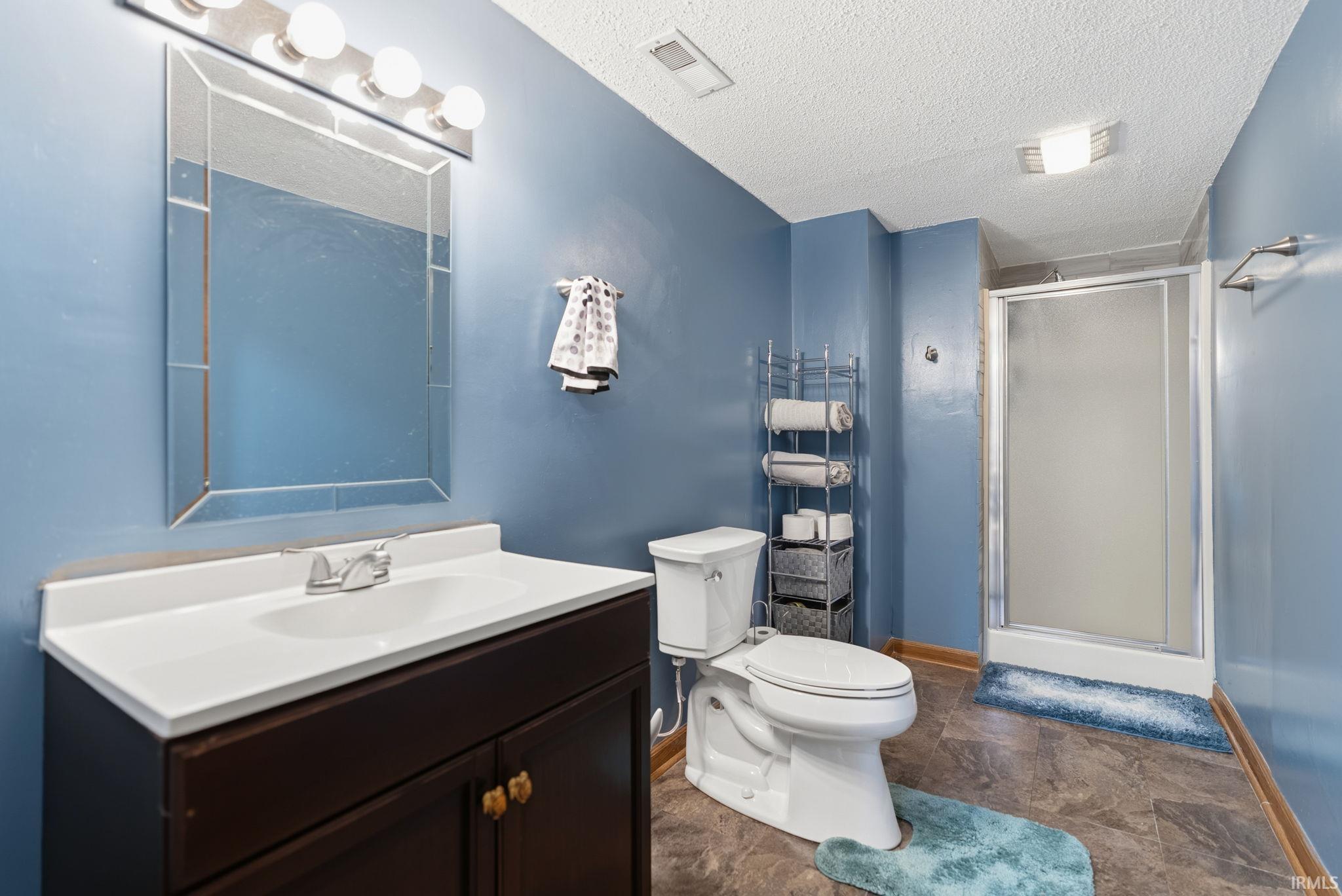 Bathroom featuring a dark wood-finish vanity with a white countertop and integrated sink, a framed mirror, a chrome light fixture, a toilet, and a stand-up shower with a frosted glass door