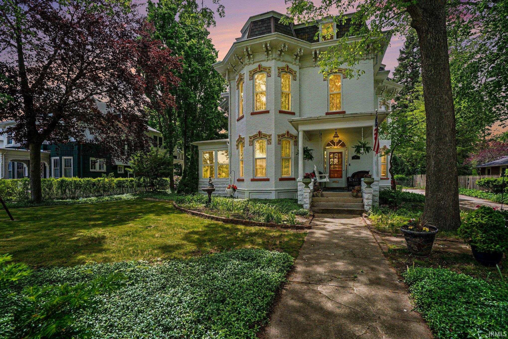 Victorian-style residence featuring a prominent front porch, intricate architectural detailing, a gabled roofline, mature trees, and a concrete pathway leading to the entrance
