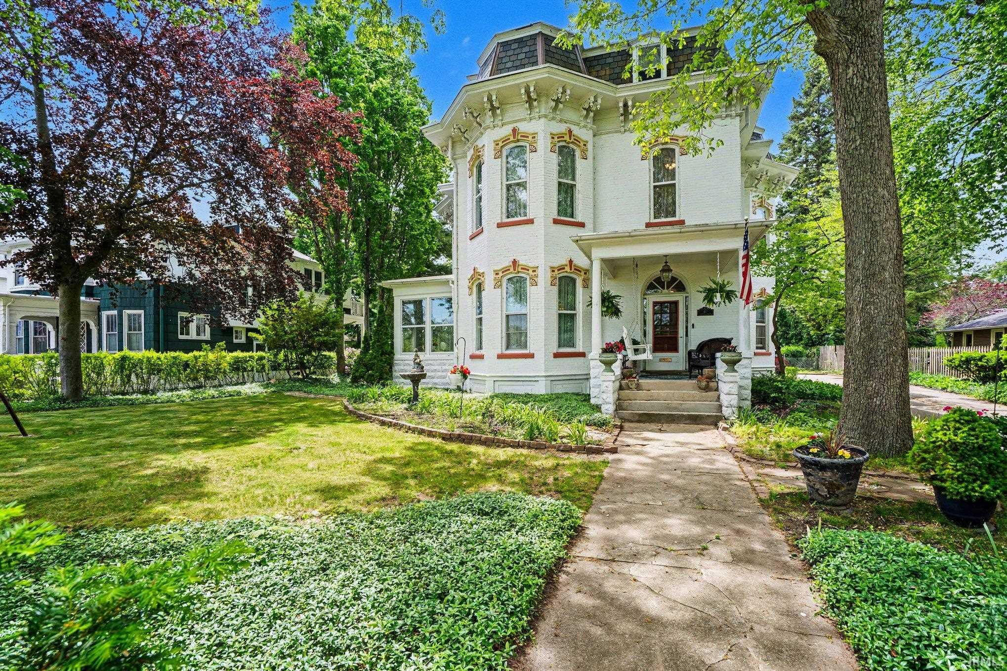 Victorian-style residence featuring a prominent front porch, intricate architectural detailing, a gabled roofline, mature trees, and a concrete pathway