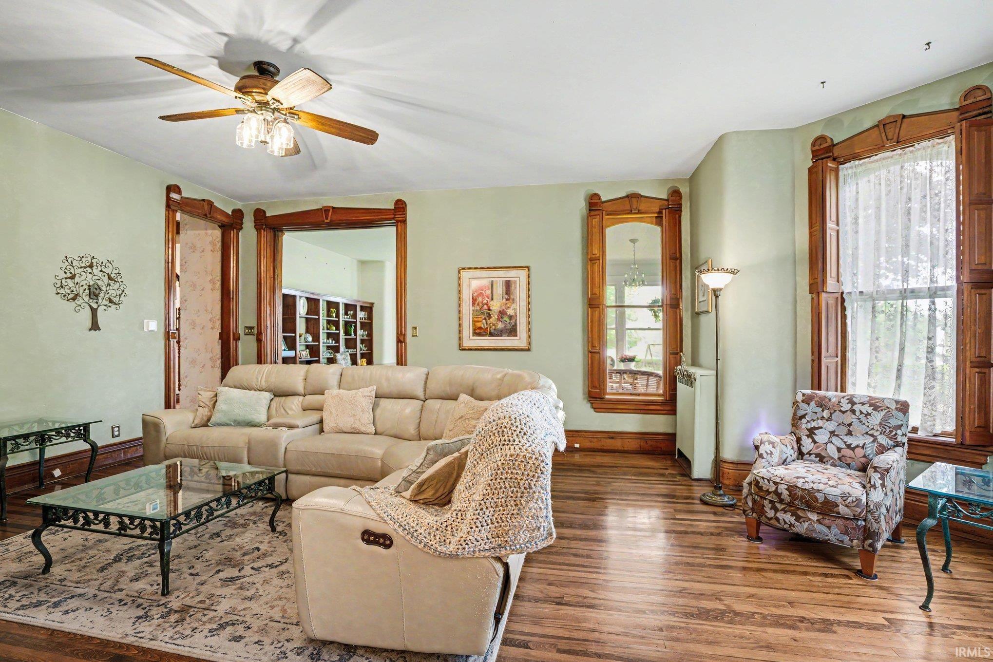 Wood-finish flooring throughout, featuring decorative wood trim around windows and doorways, a ceiling fan with integrated lighting, and multiple windows with white lace curtains