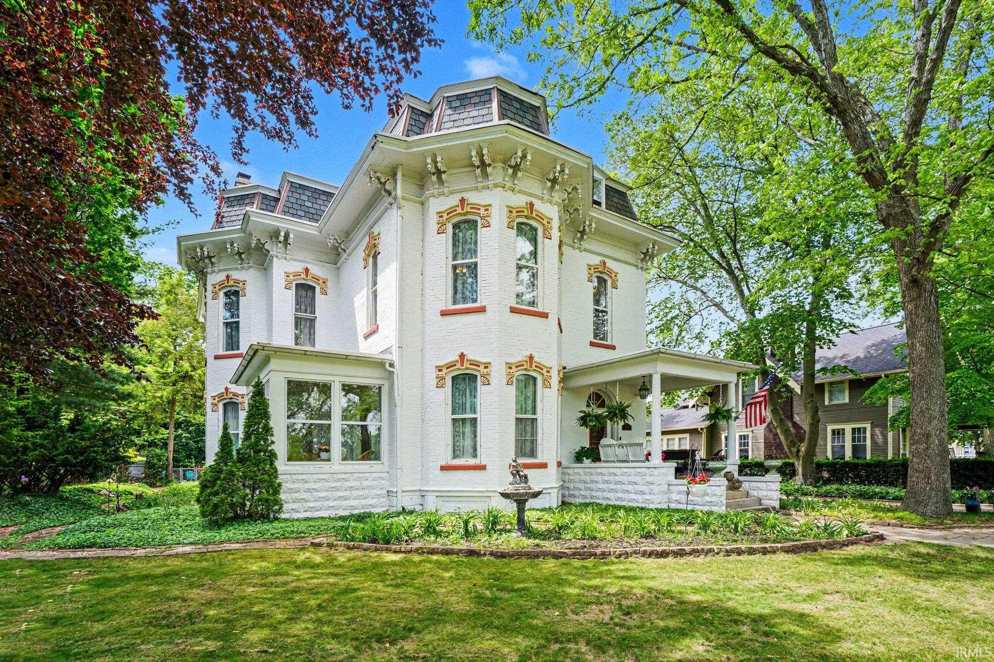 Stately Victorian residence featuring a white brick exterior, intricate decorative window trim, a multi-tiered mansard roof, a covered front porch, and mature landscaping