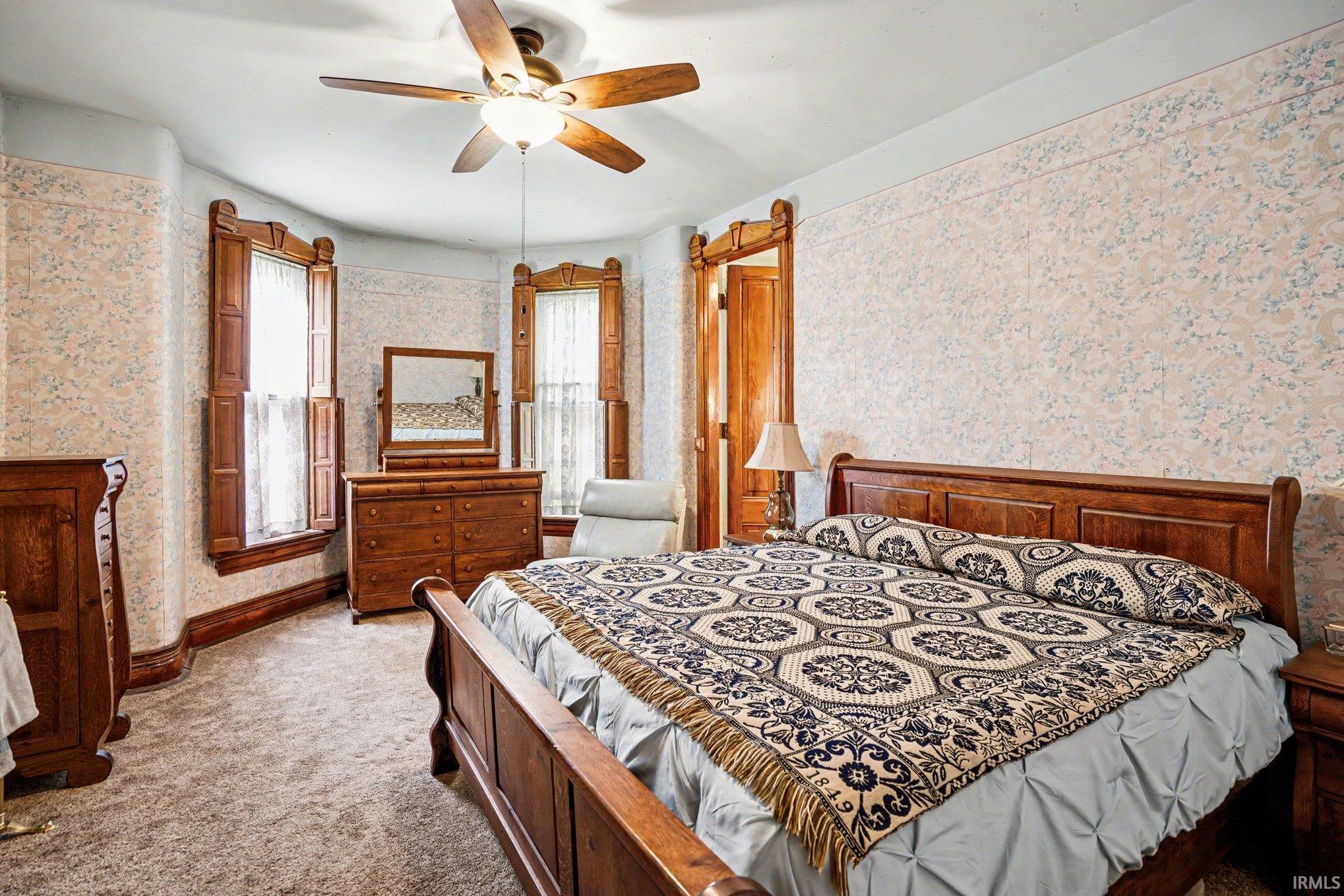 Main floor master bedroom with attached half bath with patterned wallpaper, light-colored carpeting, a ceiling fan, and bay windows featuring wood shutters