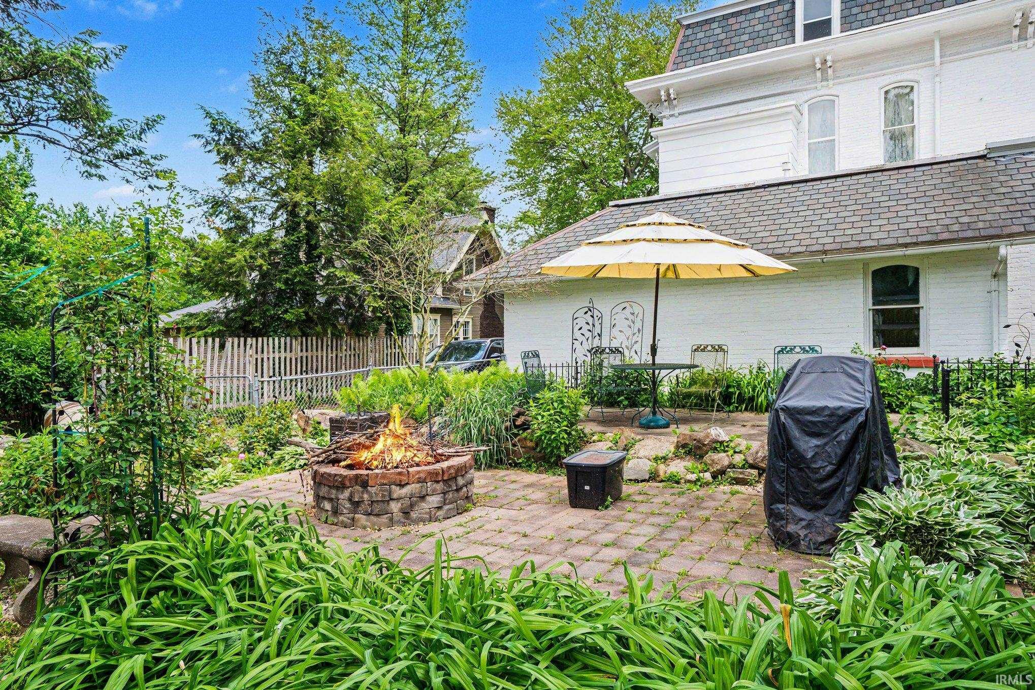 Expansive back paver patio featuring a built-in stone fire pit, lush surrounding greenery, and a white exterior facade