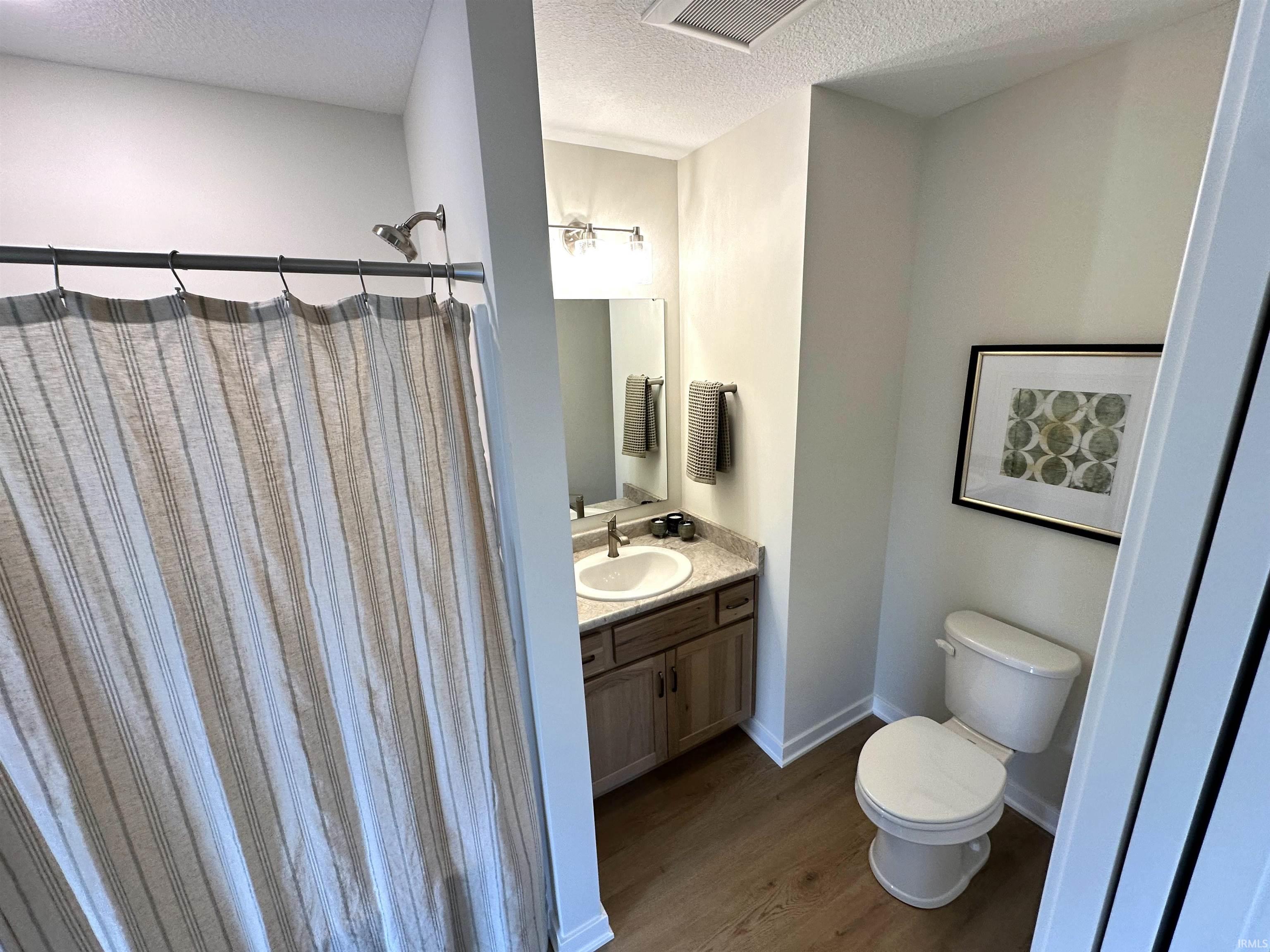 Bathroom featuring wood-finish flooring, a vanity with a light-toned countertop and integrated sink, a wall-mounted mirror, and a shower with a chrome fixture