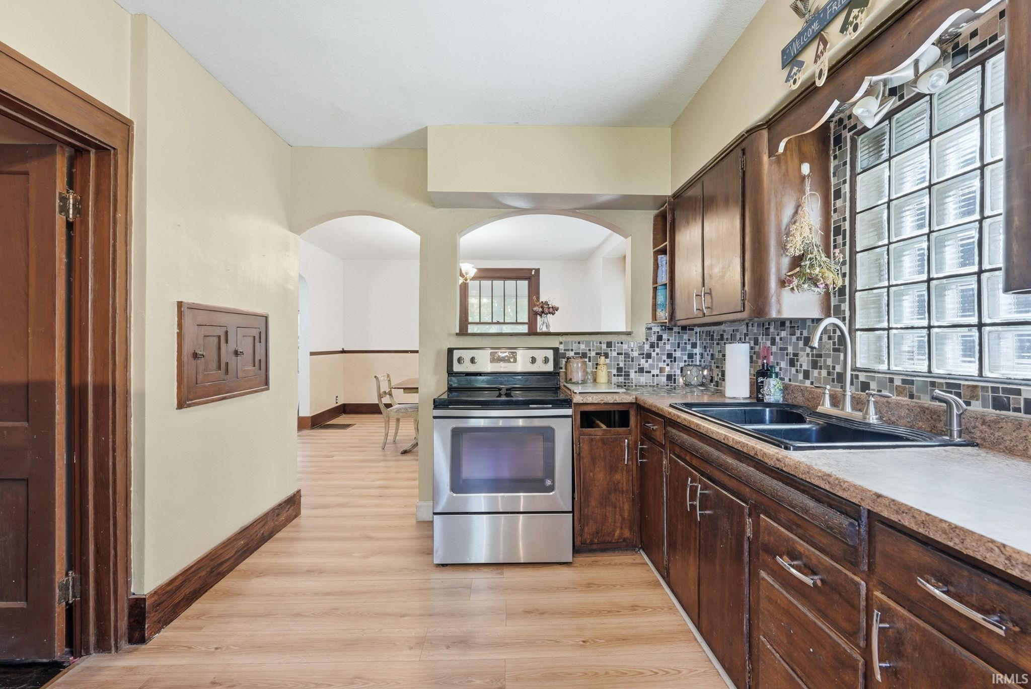 Kitchen featuring wood-finish flooring, dark wood cabinetry, a mosaic tile backsplash, and a glass block window