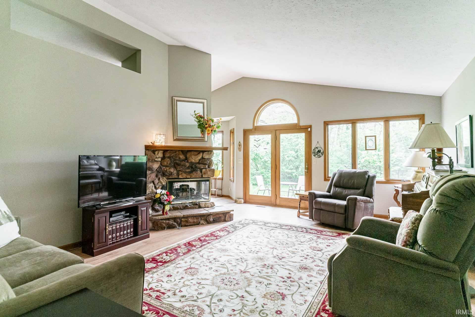 Spacious living area featuring a stone-clad fireplace with hearth, wood-finish flooring, vaulted ceilings, and French doors with an arched transom