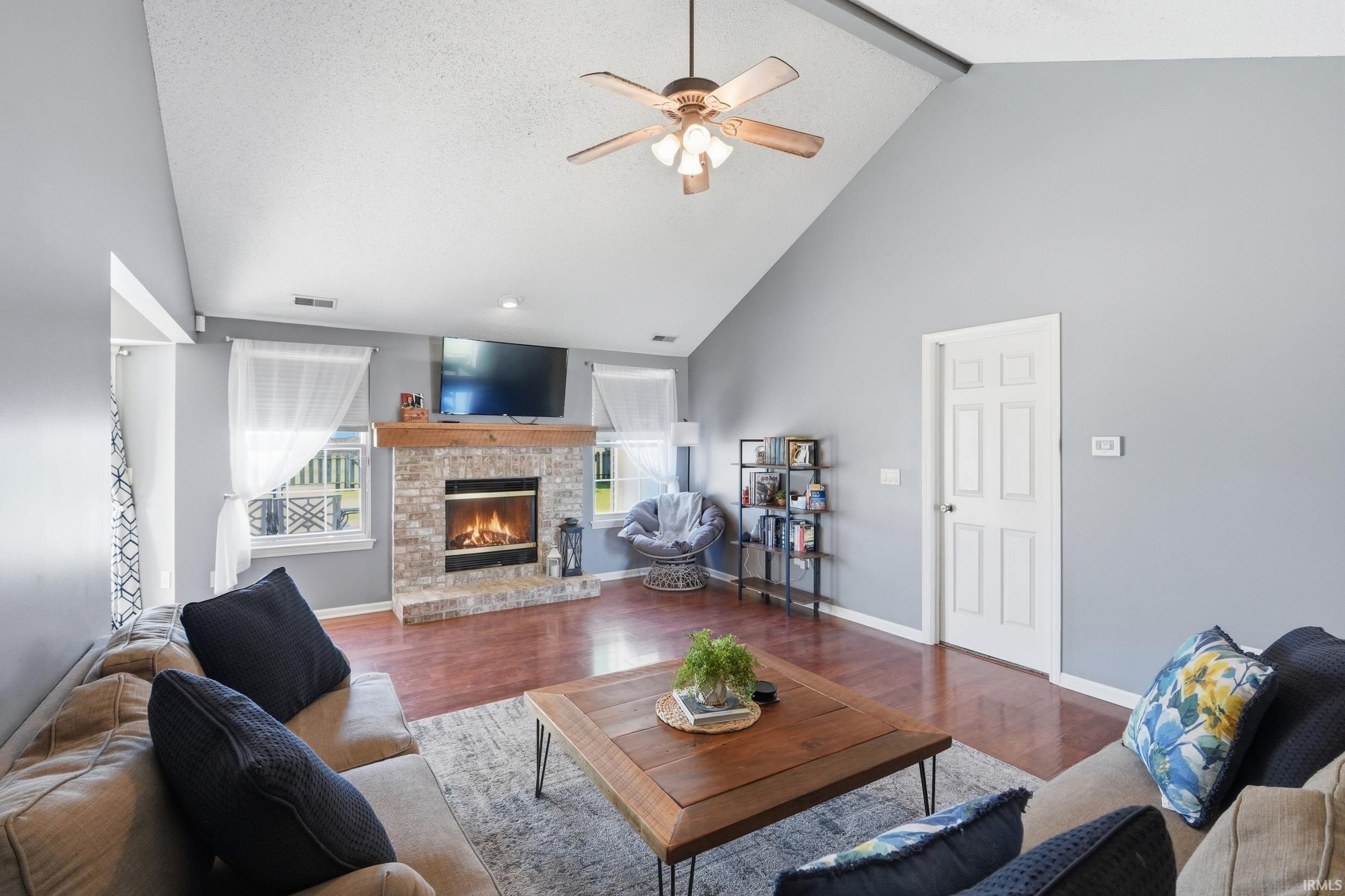 Living area featuring a vaulted ceiling with a ceiling fan, a stone-front fireplace with a wood mantle, wood-finish flooring, and multiple windows