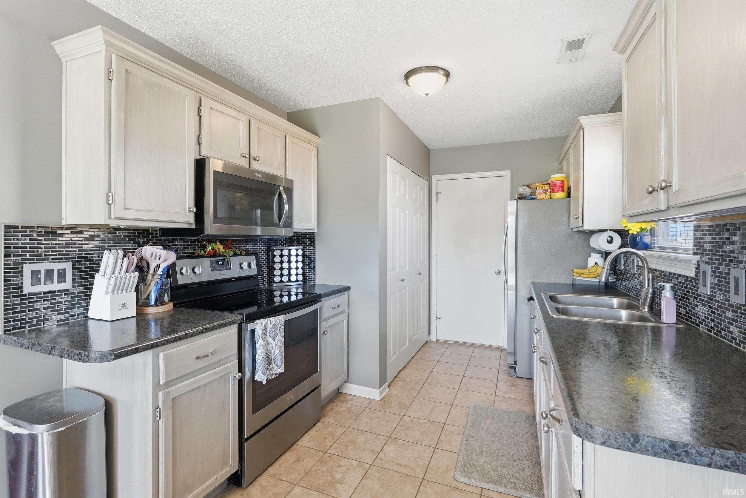 Kitchen featuring wood-finish cabinetry, black countertops, stainless steel appliances, mosaic tile backsplash, and tile flooring