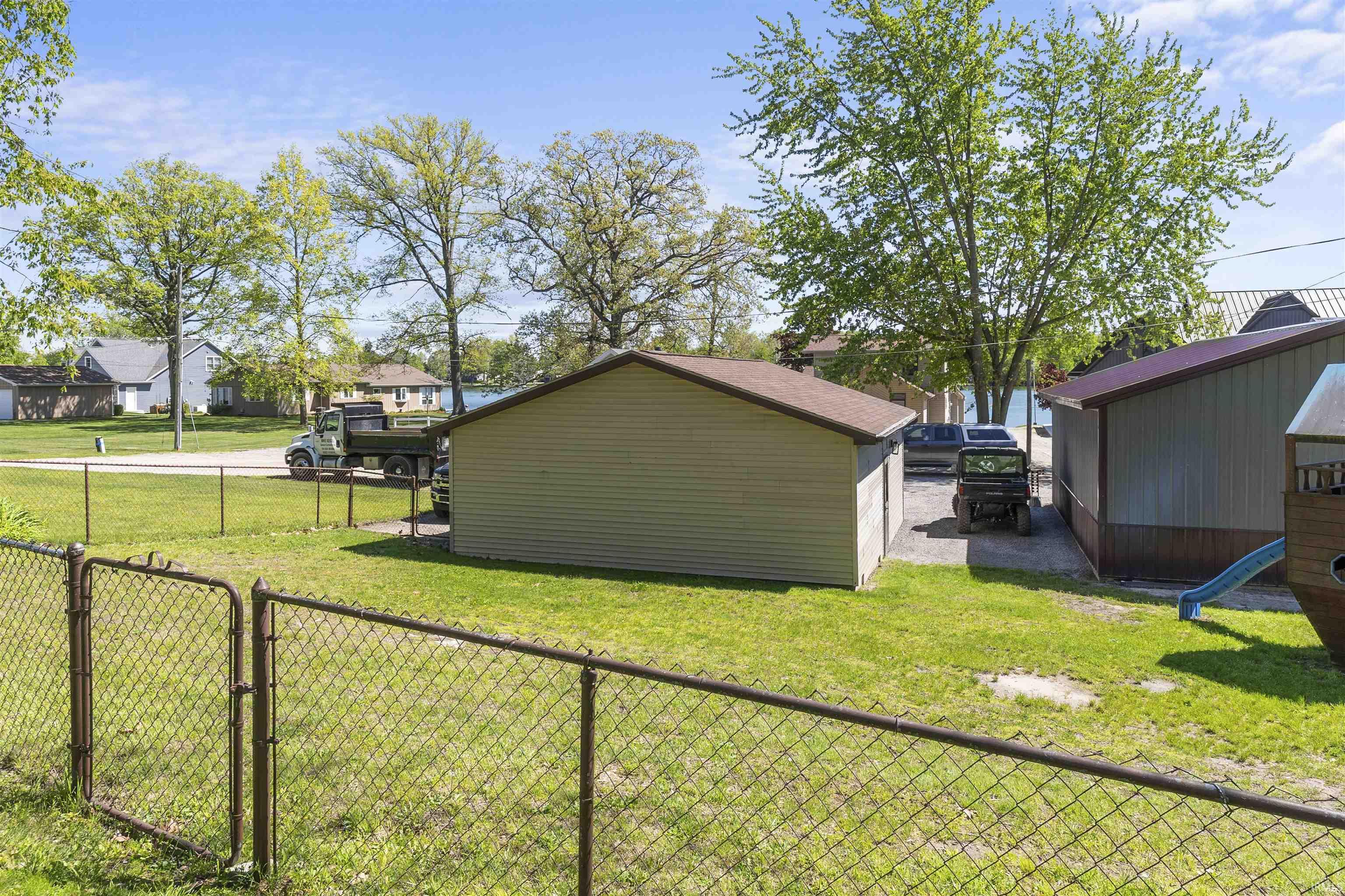 Expansive grass lot featuring a chain-link fence, two outbuildings with sloped roofs, and a paved driveway