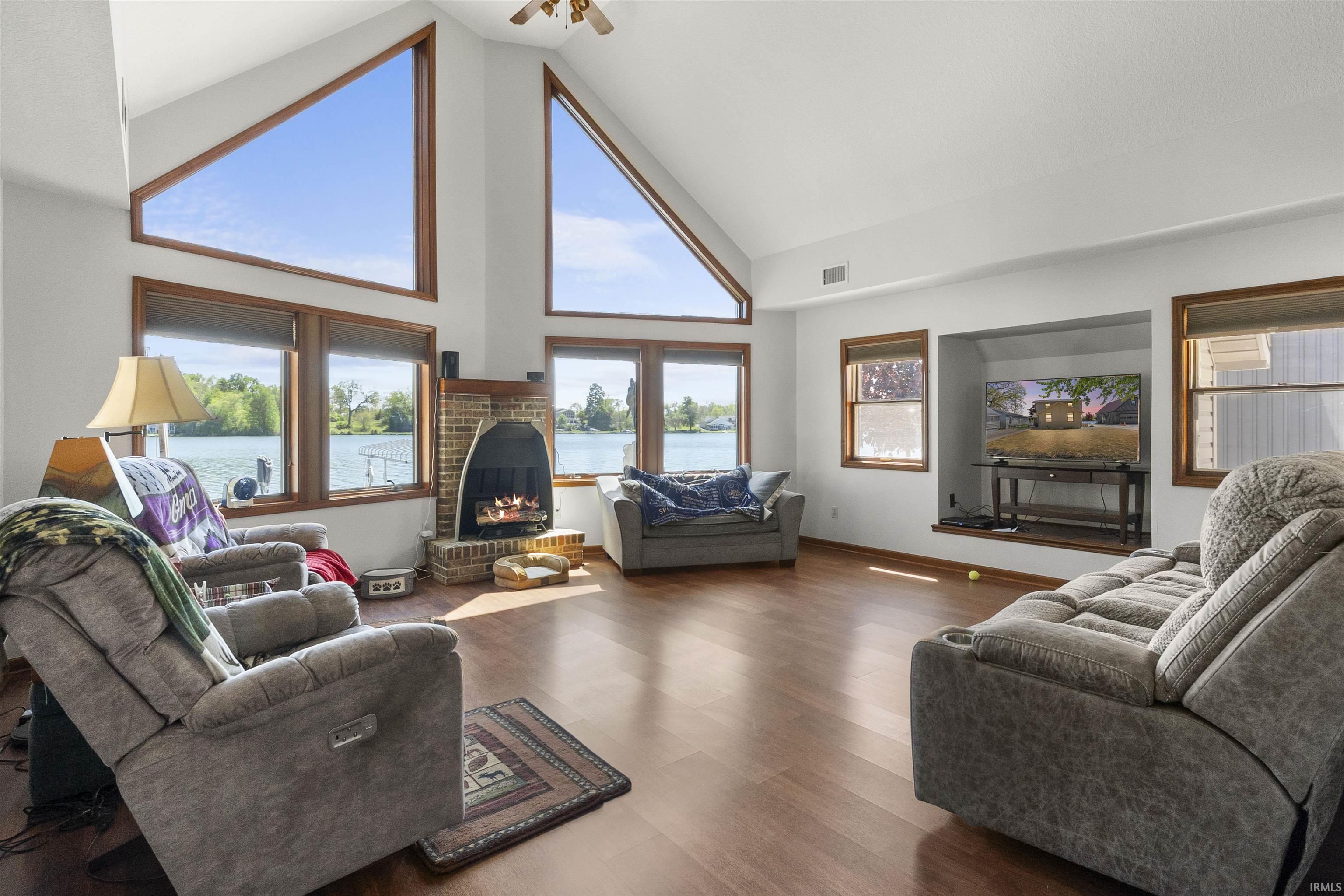 Great room featuring vaulted ceilings, wood-finish flooring, a brick fireplace, and expansive windows showcasing a waterfront setting