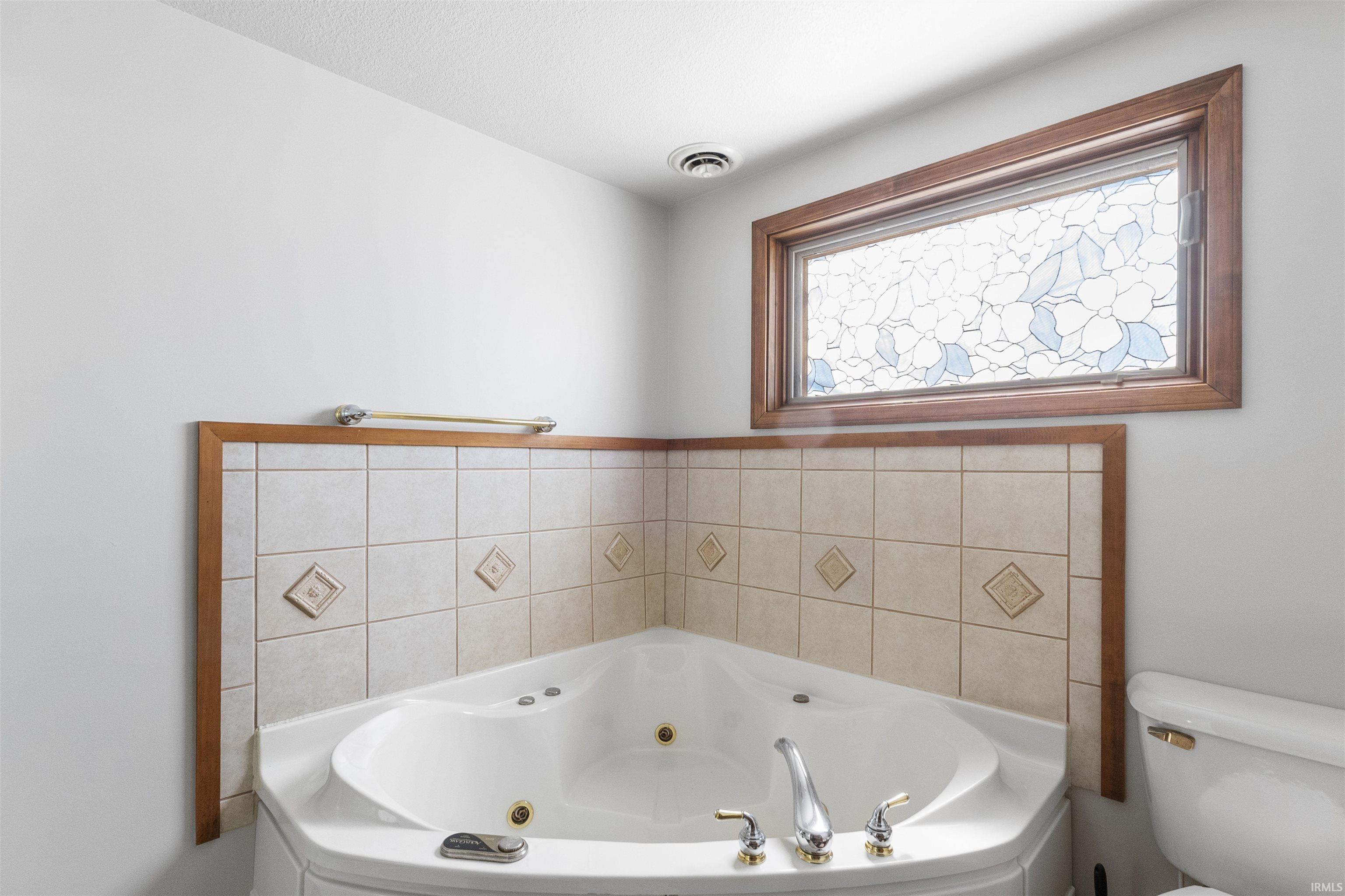 Corner jetted tub with tile surround and wood-frame window featuring a decorative glass insert