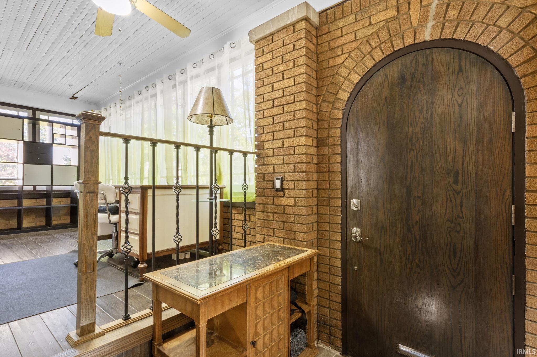 Arched entry door set within a brick wall, featuring dark wood-finish flooring, a white plank ceiling, and an ornate metal railing