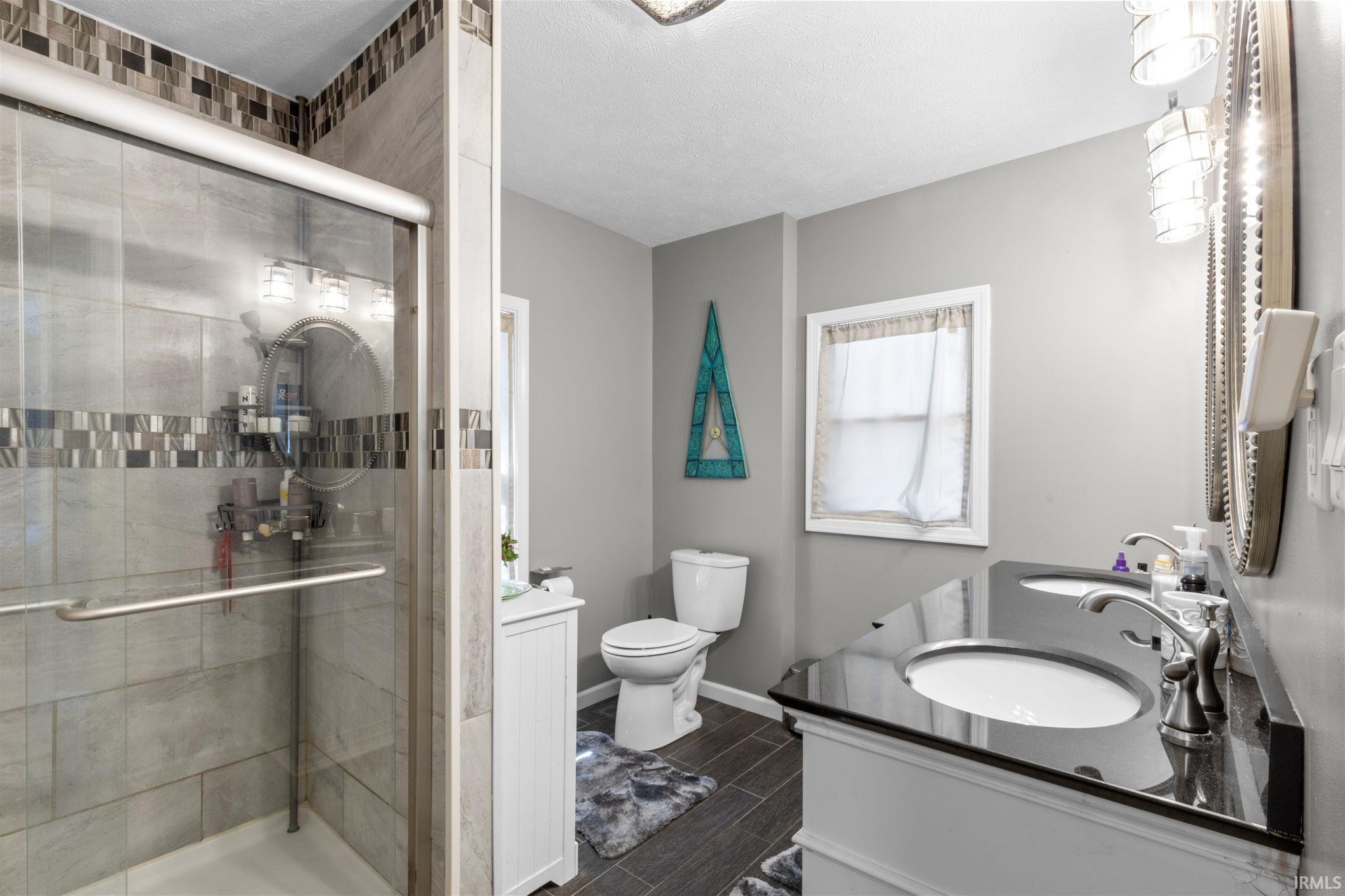 Bathroom featuring a glass-enclosed shower with tile surround, dual vanity with dark countertops, wood-finish flooring, and a window