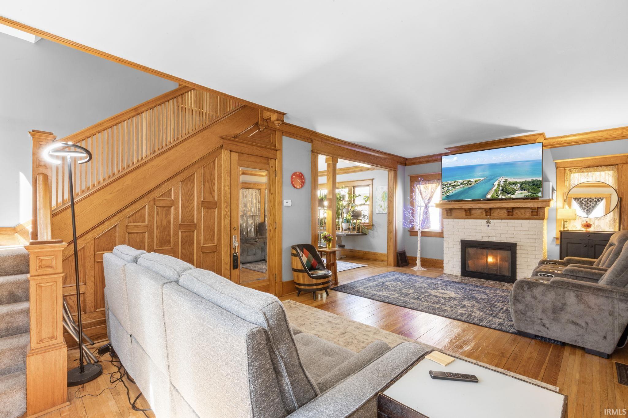 Living area featuring wood-finish flooring, a fireplace with a white brick surround and wood mantel, and a prominent wood staircase with balusters