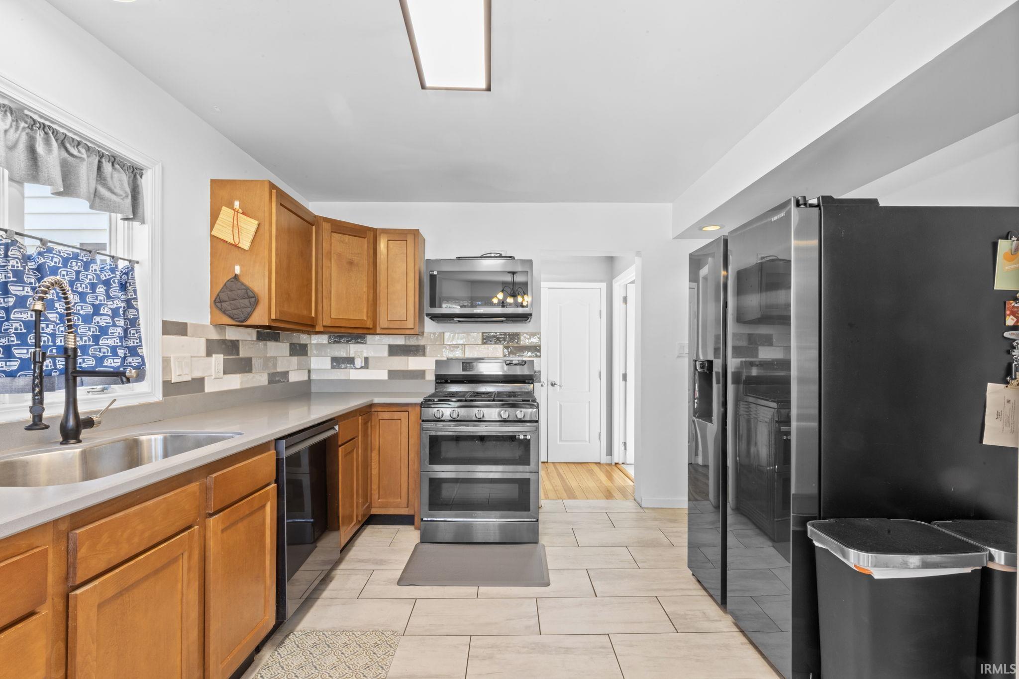 Kitchen featuring wood cabinetry, light-toned countertops, a stainless steel sink with a black pull-down faucet, a patterned tile backsplash, and light-colored tile flooring